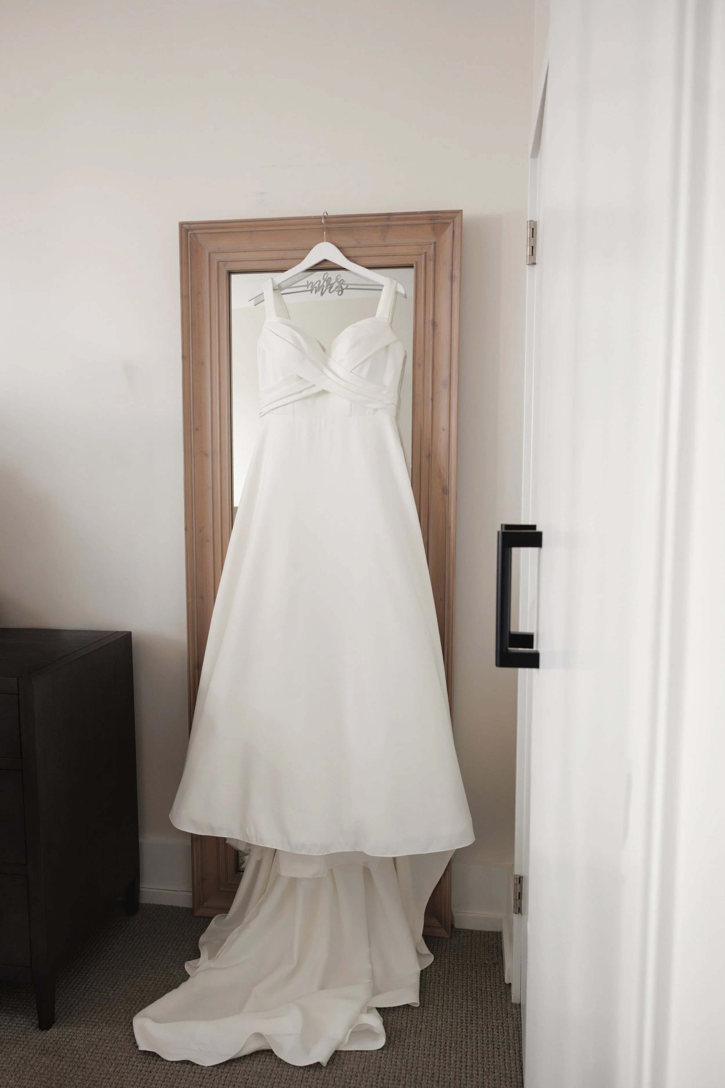 A white wedding dress hanging on a hanger attached to a mirror with a wooden frame.