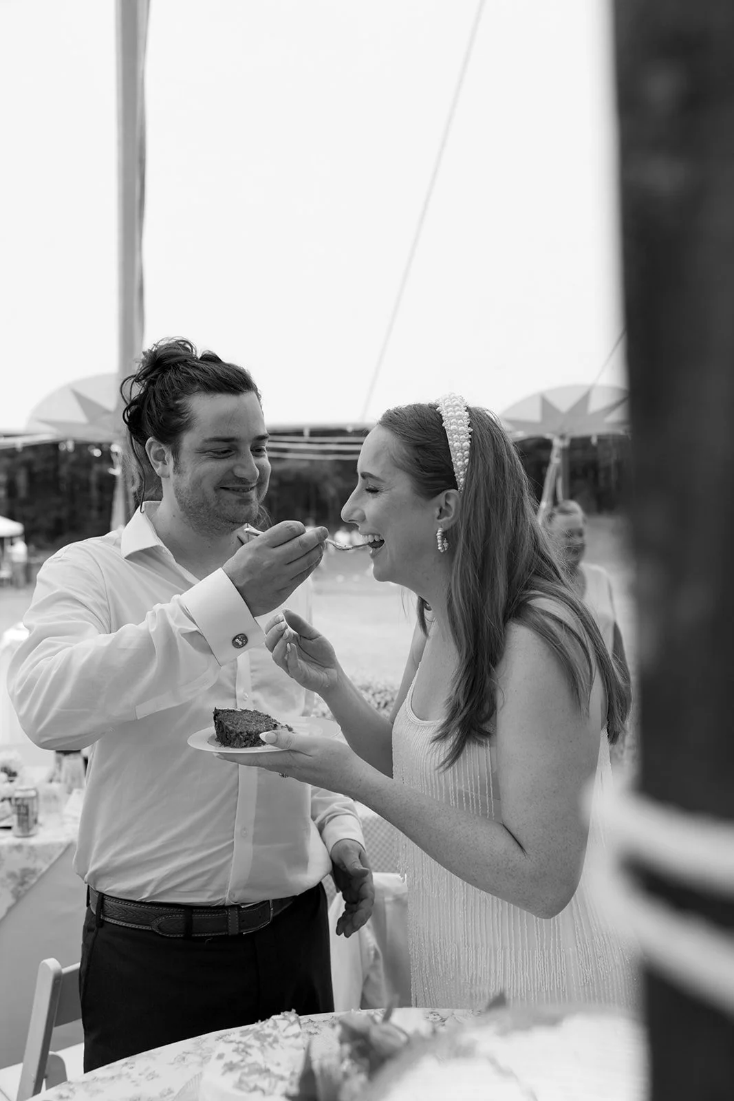 A joyful couple sharing a cake at an outdoor celebration, possibly a wedding, with the man feeding the woman a slice.