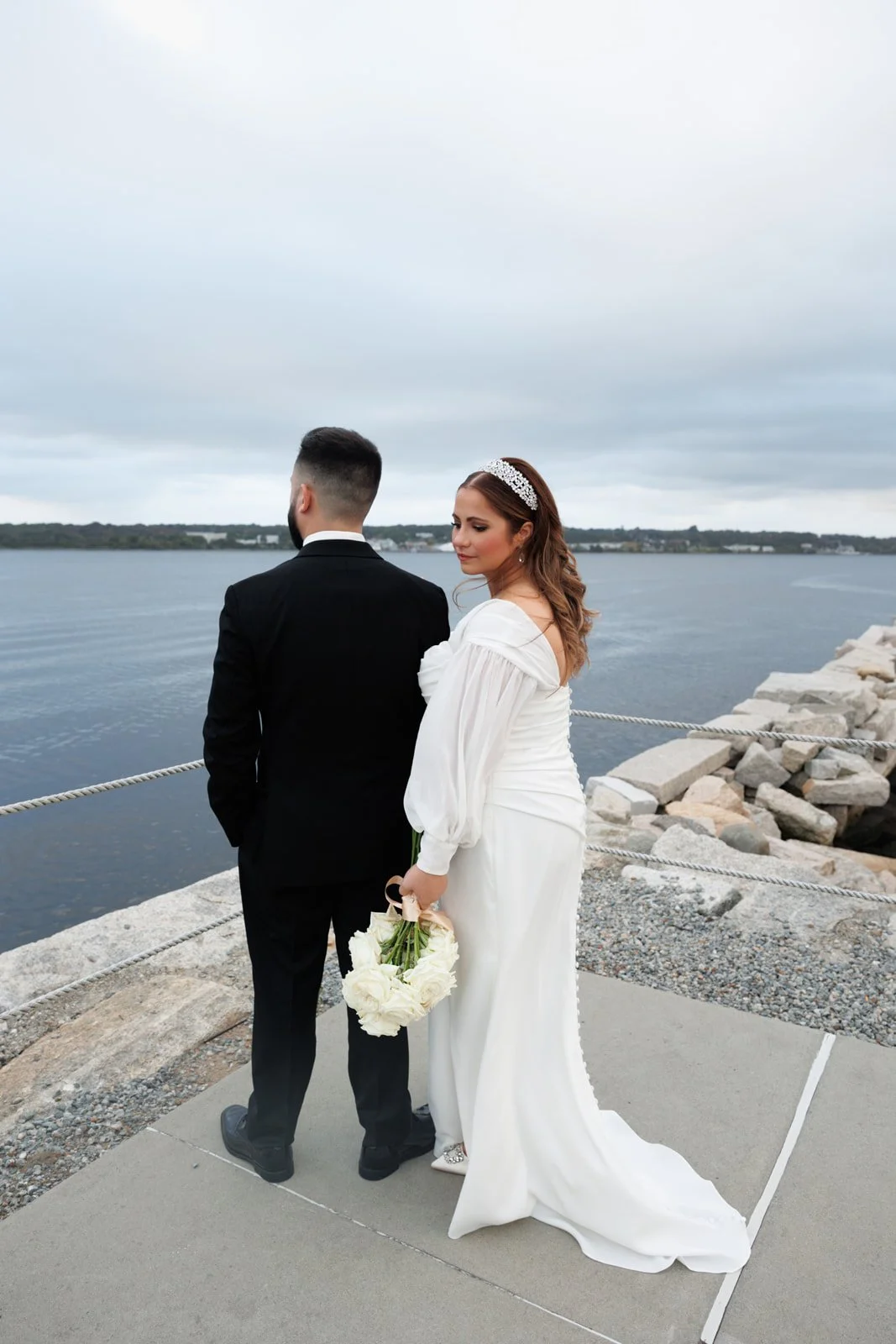 Bride and groom standing by a waterfront, with the bride holding a bouquet of white roses and wearing a white wedding dress and tiara, seen from the back and side in an outdoor setting.