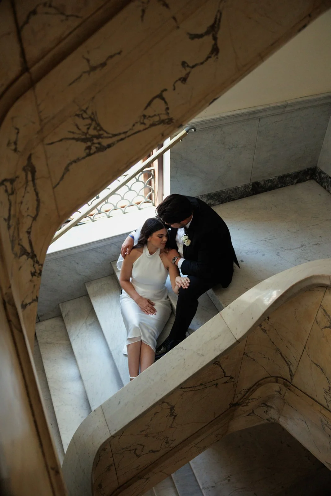 A bride and groom sitting together on a marble staircase, viewed from above through a rounded opening, with the bride in a white dress and the groom in a black suit.