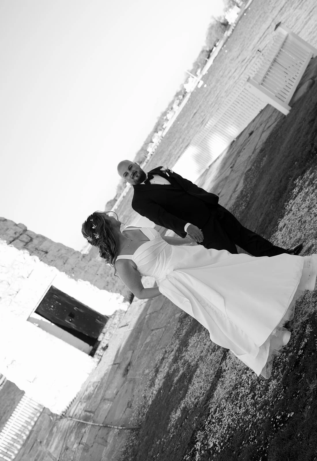 A bride and groom holding hands, walking together outdoors near a stone building and a body of water.