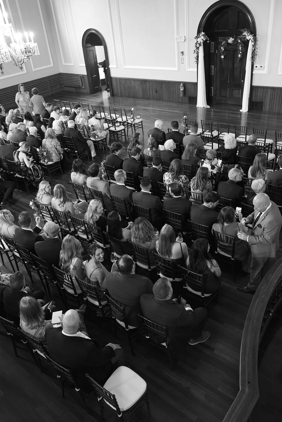 People attending a wedding ceremony in an elegant hall, seated in rows facing a decorated ornate door with floral arrangements, some guests taking photos or holding drinks.