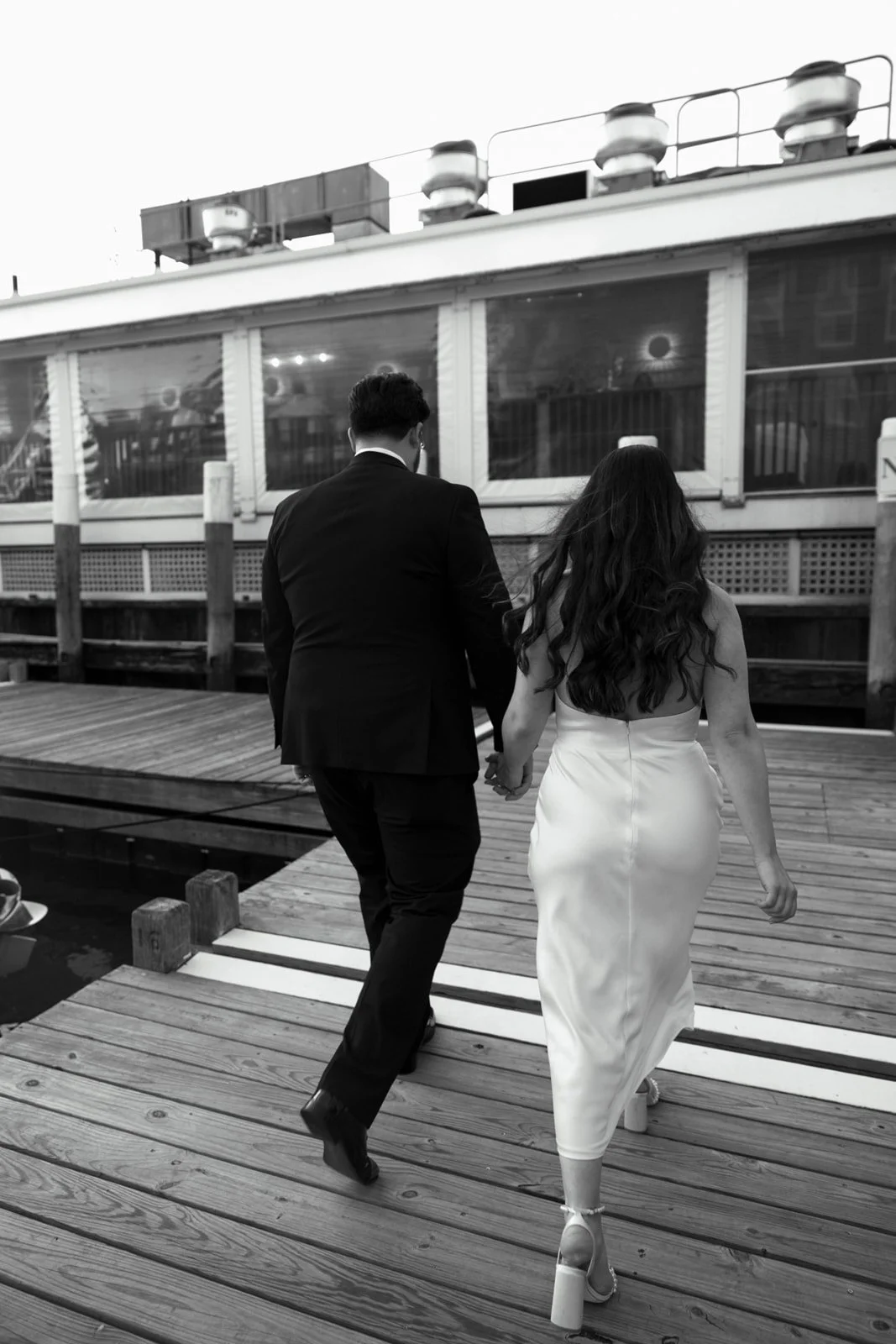 A couple dressed in formal attire, walking hand in hand on a wooden dock beside a boat, with the woman in a white dress and high heels and the man in a black suit, in an outdoor setting.