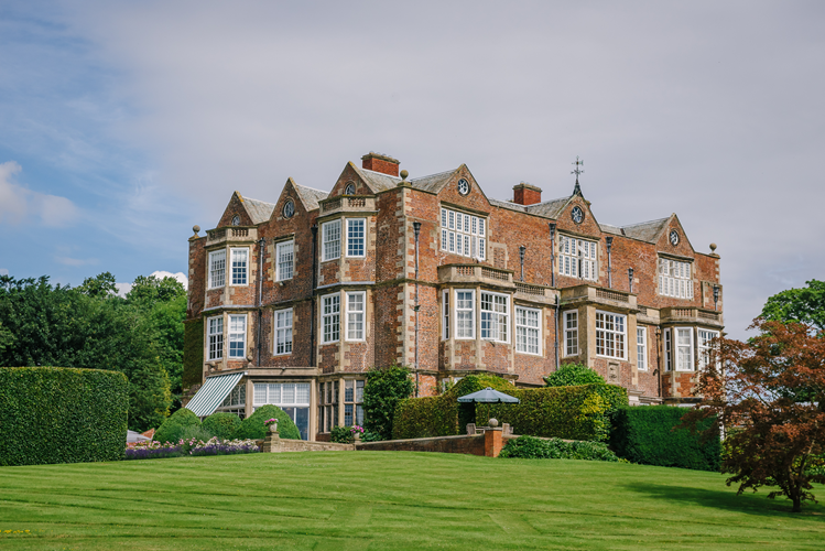 A large, historic brick mansion with multiple stories, numerous large windows, and a well-maintained lawn and garden in the foreground.