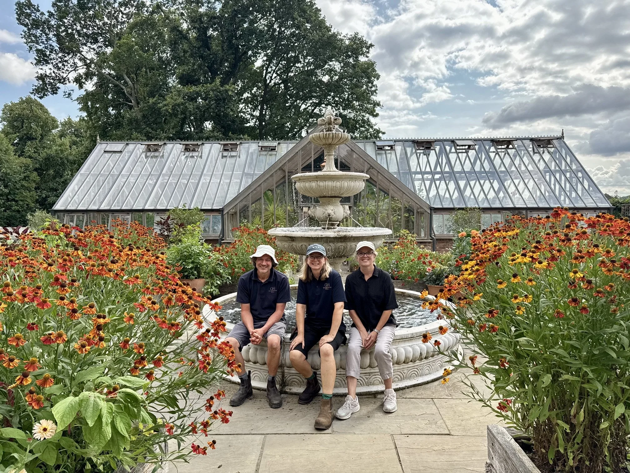 Image shows a man and two women sitting on the edge of a fountain in a kitchen garden with a large glasshouse behind them