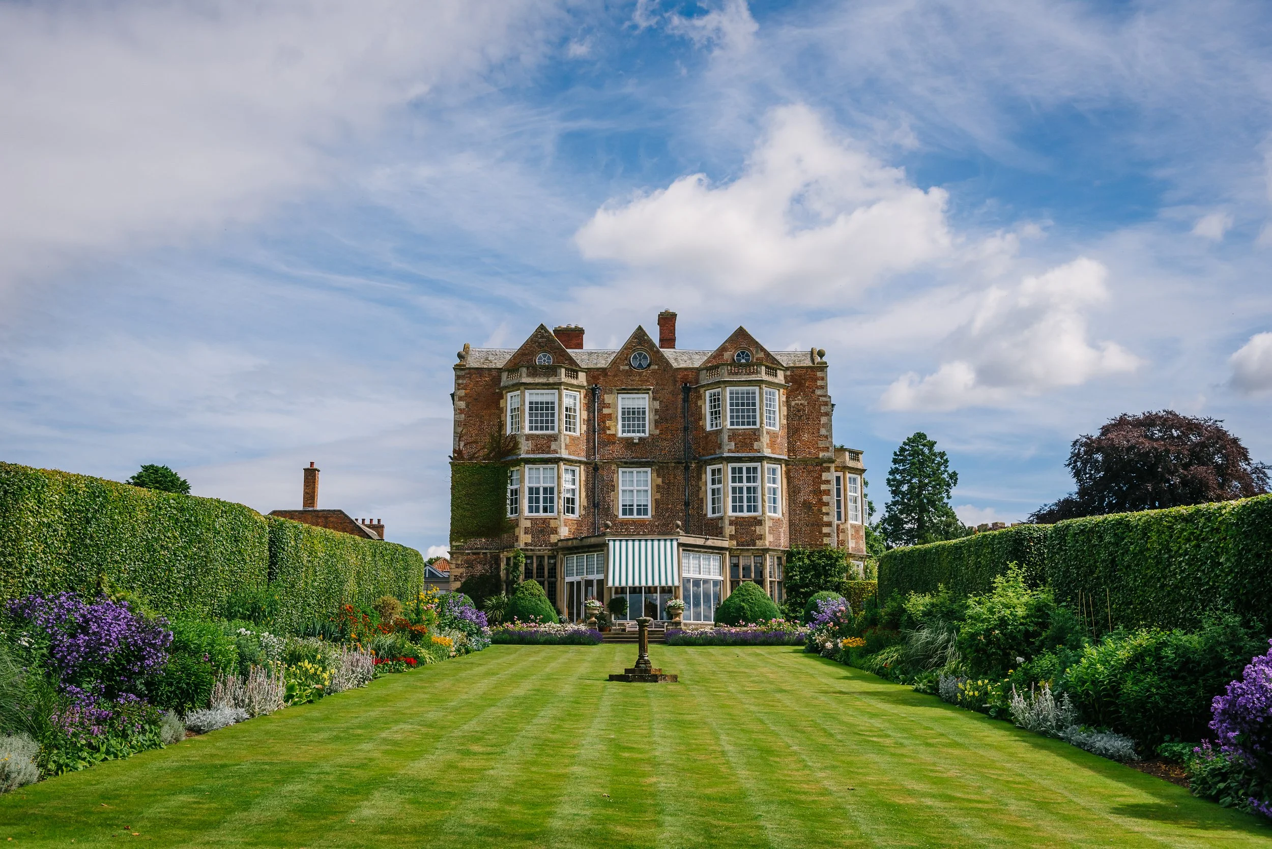 Photograph of a stately home and gardens, some green lawn and two borders lined with a beech hedge with the house in the background