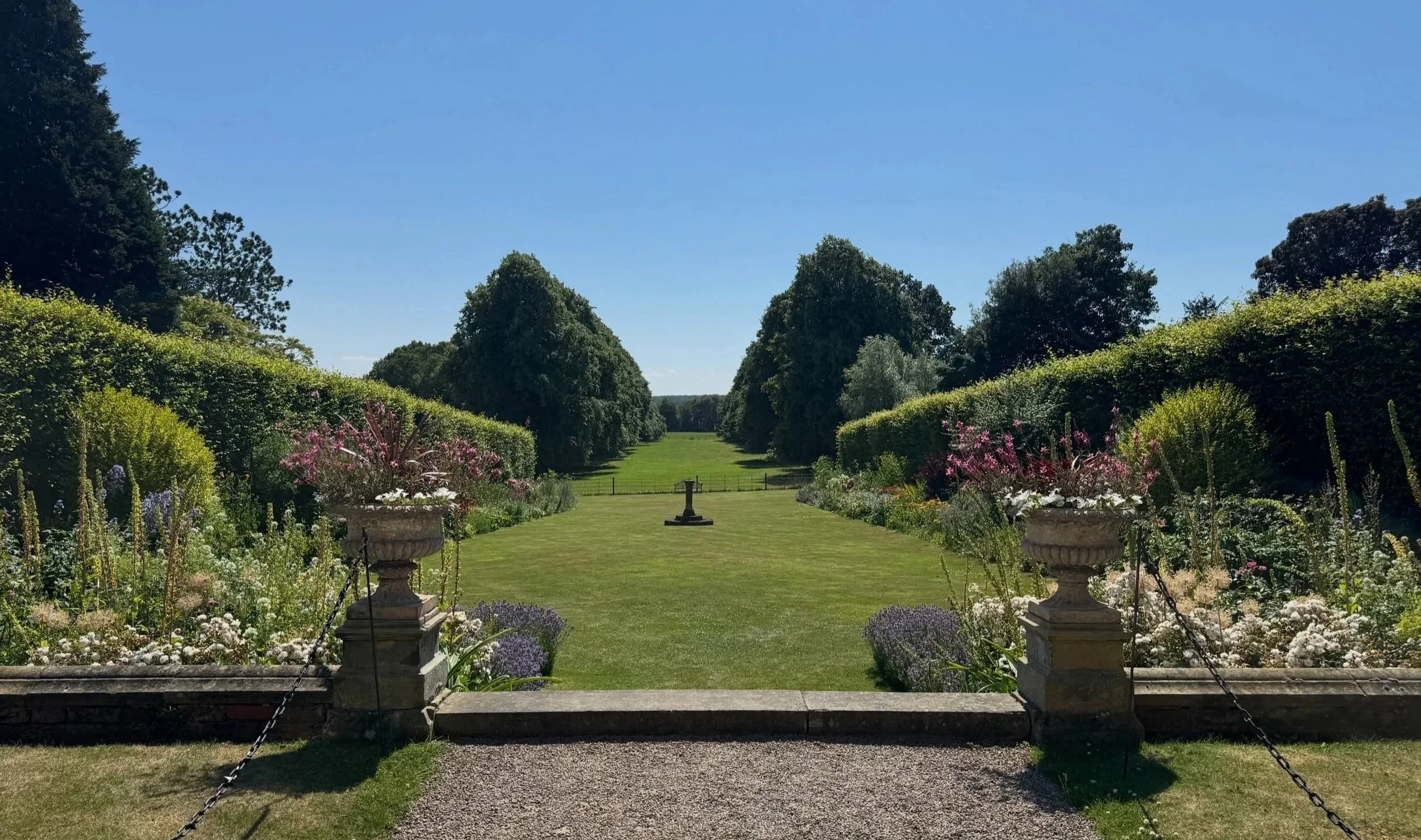 Image shows two garden urns in the foreground lawn of grass, two herbaceous borders either side, two beech hedges flanking them and in the distance a Lime tree walk - from the gardens at Goldsborough Hall, North Yorkshire