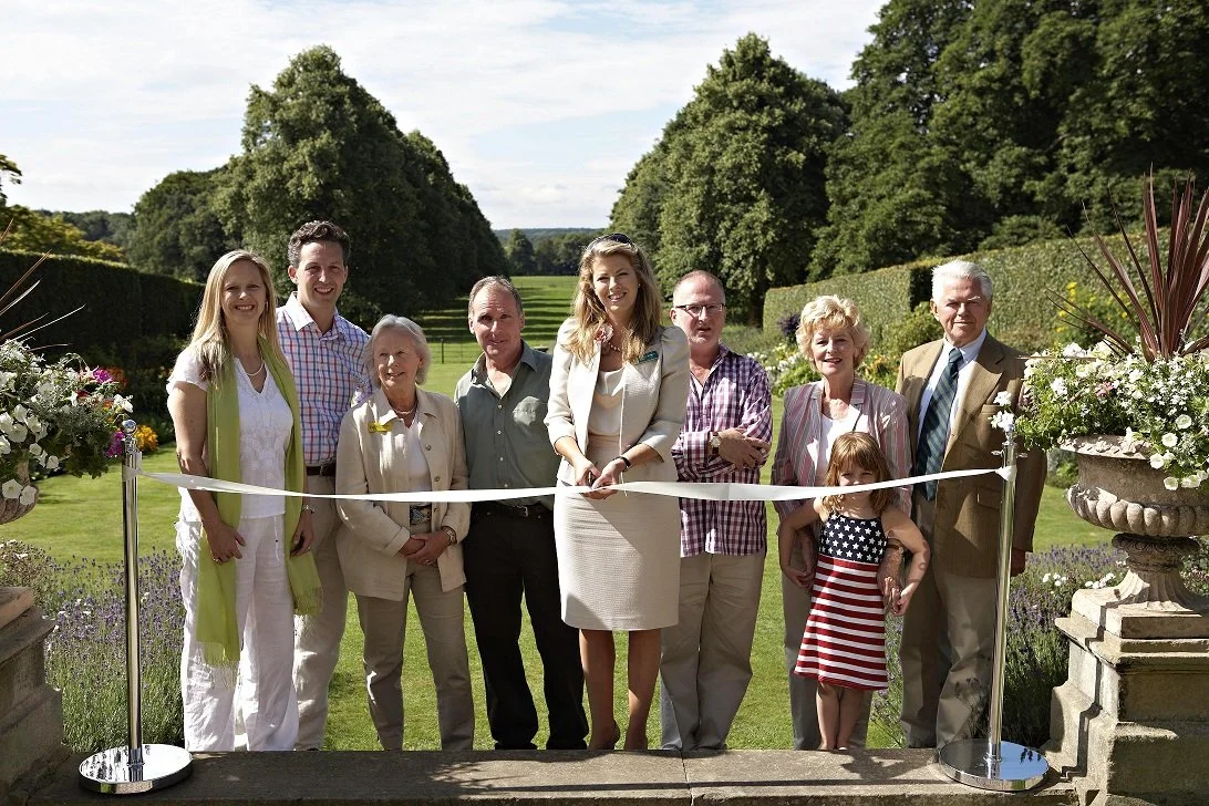 A woman cutting a ribbon to show that Goldsborough gardens were re-opening. She is standing in the gardens with people around her
