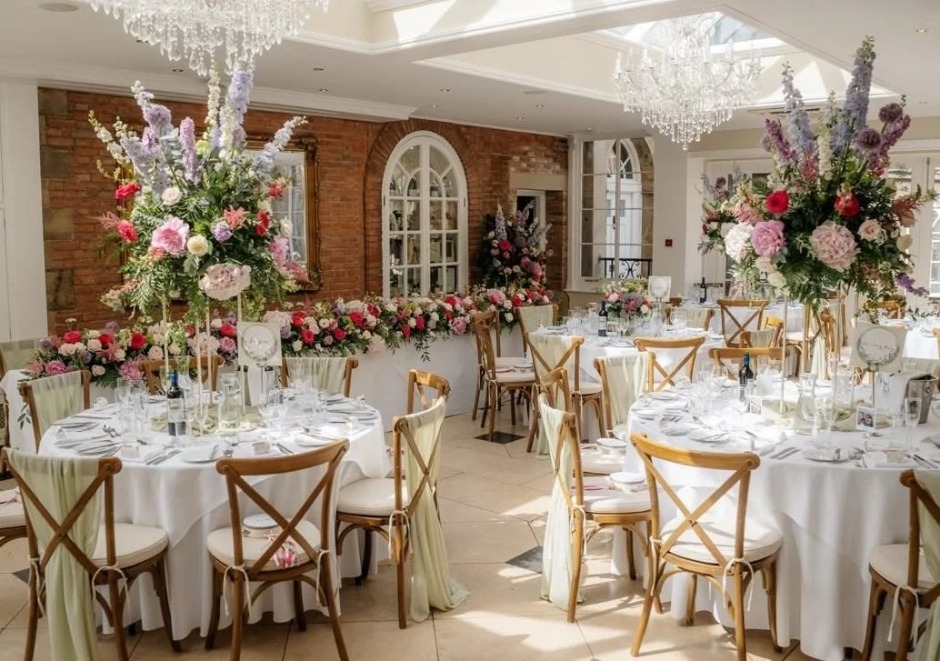 Image shows a wedding breakfast in the Orangery at Goldsborough Hall, North Yorkshire with round and long tables, white linen, wooden crossback chairs and stunning large floral arrangements in blues and pinks