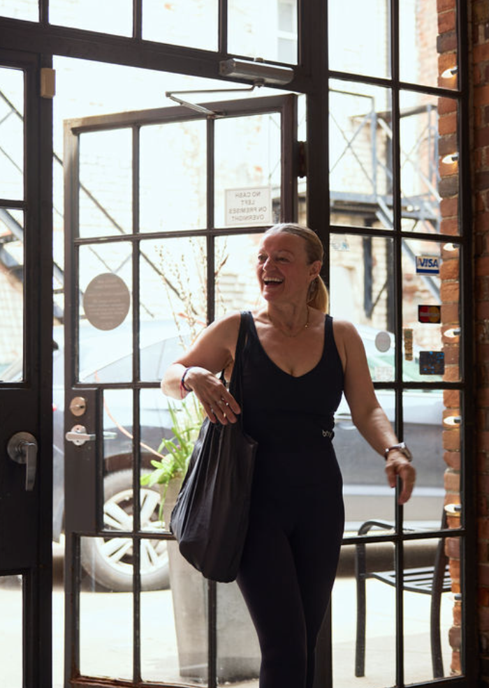 A woman in black clothing standing at an open glass door, smiling, with a bag over her shoulder inside a brick-walled building.