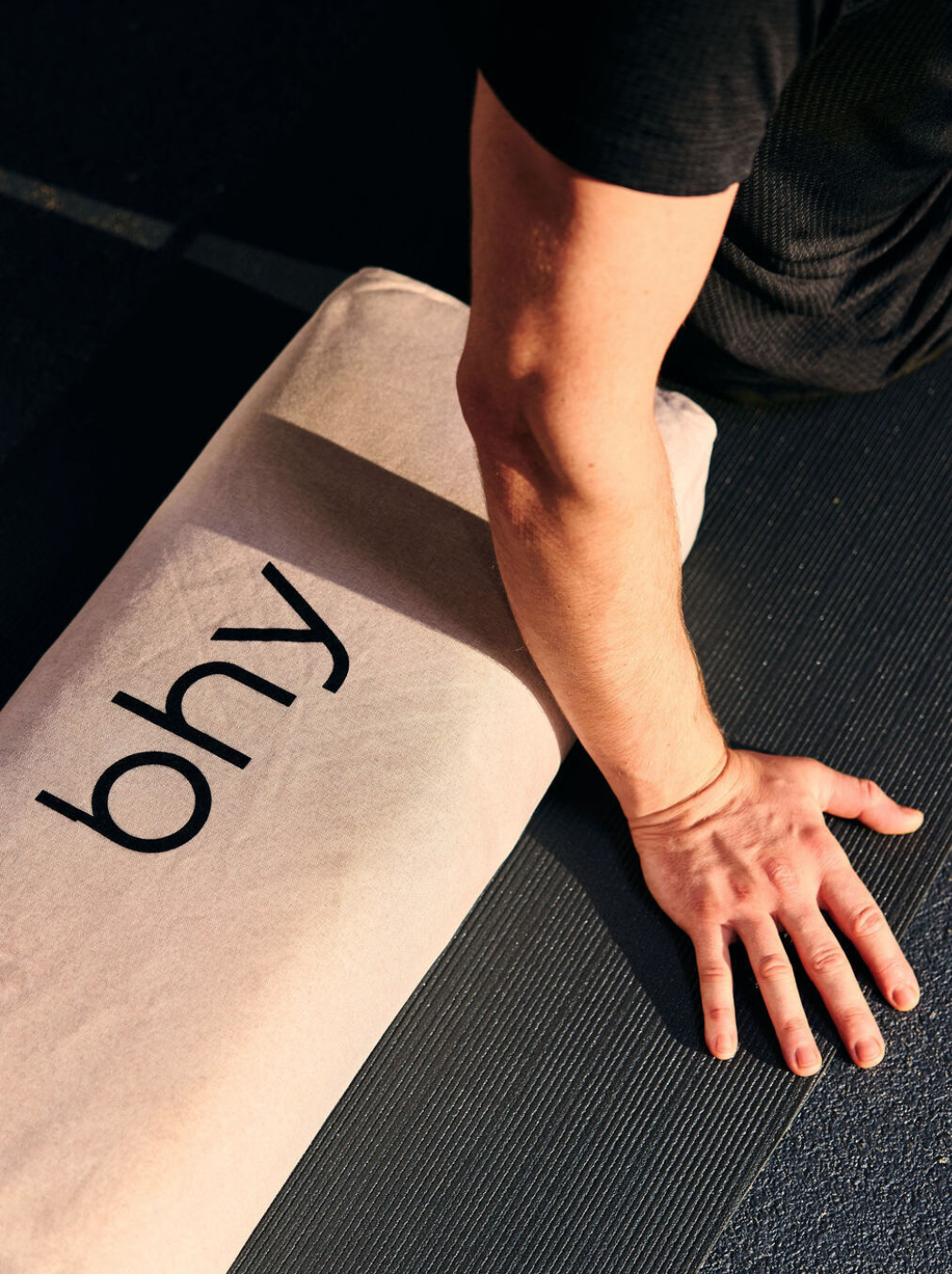 A person in black clothing is performing a push-up on a light-colored exercise mat with the word 'bhy' printed on it, on a dark exercise surface.