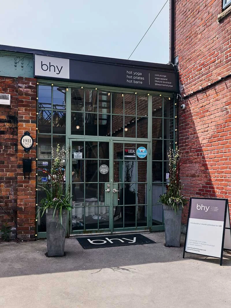 Exterior view of a brick building entrance with glass doors, signage for 'bhy' wellness studio offering hot yoga, hot pilates, and hot barre, and outdoor planters with flowers.
