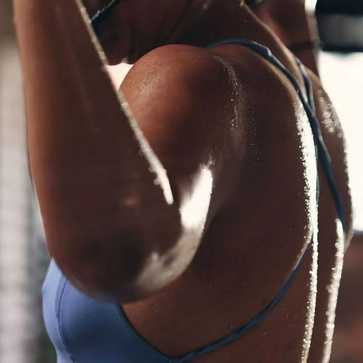 Close-up of a woman's shoulder and upper arm, with sunlight shining on her skin in a fitness or athletic setting.