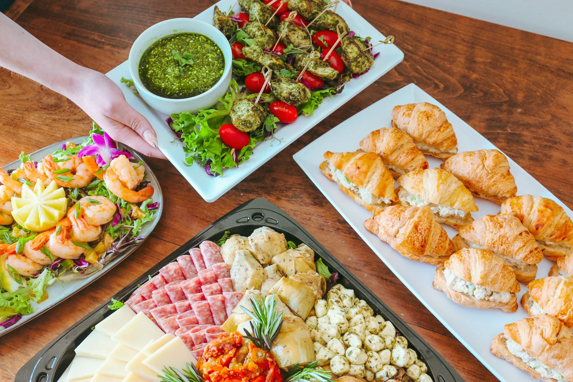 A spread including grilled pesto skewers, chicken salad croissants, an antipasto tray, and steamed shrimp
