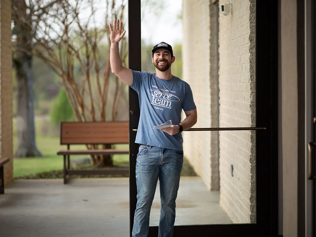 A man with a beard wearing a cap and blue t-shirt waves from a door opening, holding papers in his left hand, with a bench and trees outside in the background.