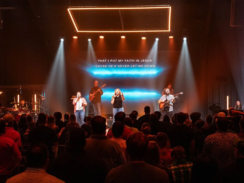 A church service with a band performing on stage, including singers, guitarists, and a drummer, with a large screen displaying song lyrics and an audience watching.