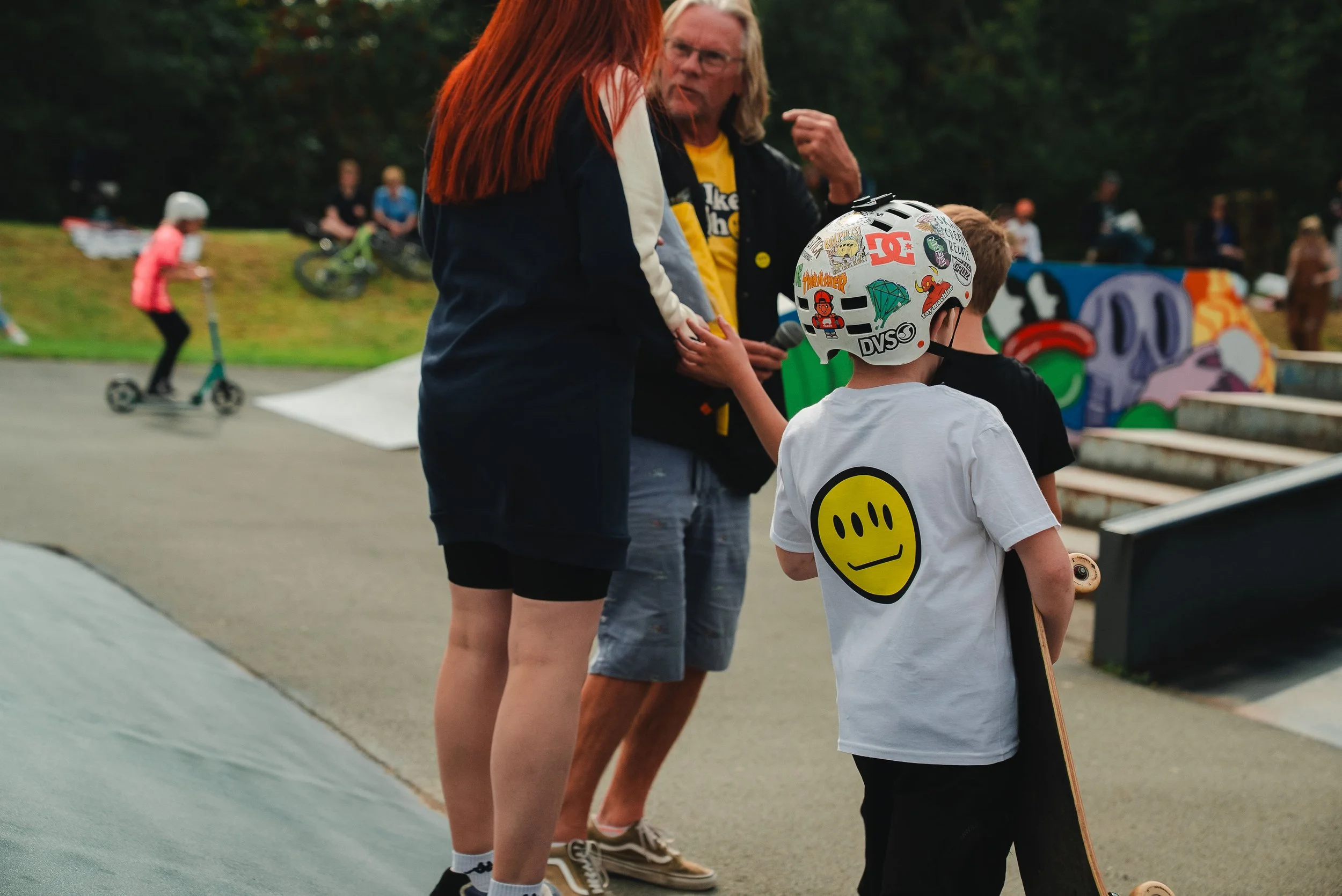Live Like Ralph A group of people at a skate park, with a child holding a skateboard, two women, and a man talking to the child. In the background, other children are riding scooters and bikes, and there are colorful graffiti murals on the ramps.