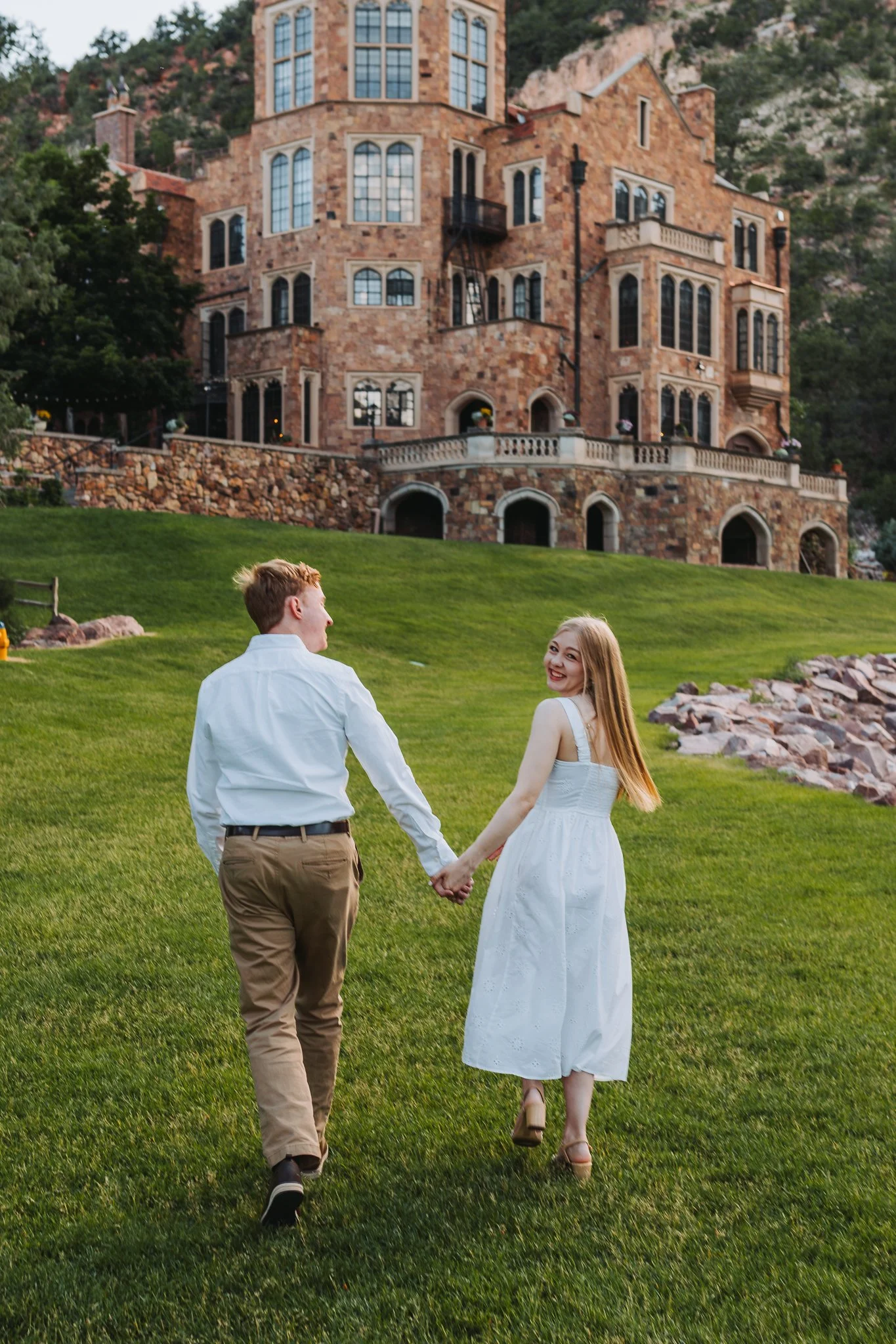 A young couple dressed in white holding hands and walking on a grassy hill, with a large, ornate brick mansion in the background.