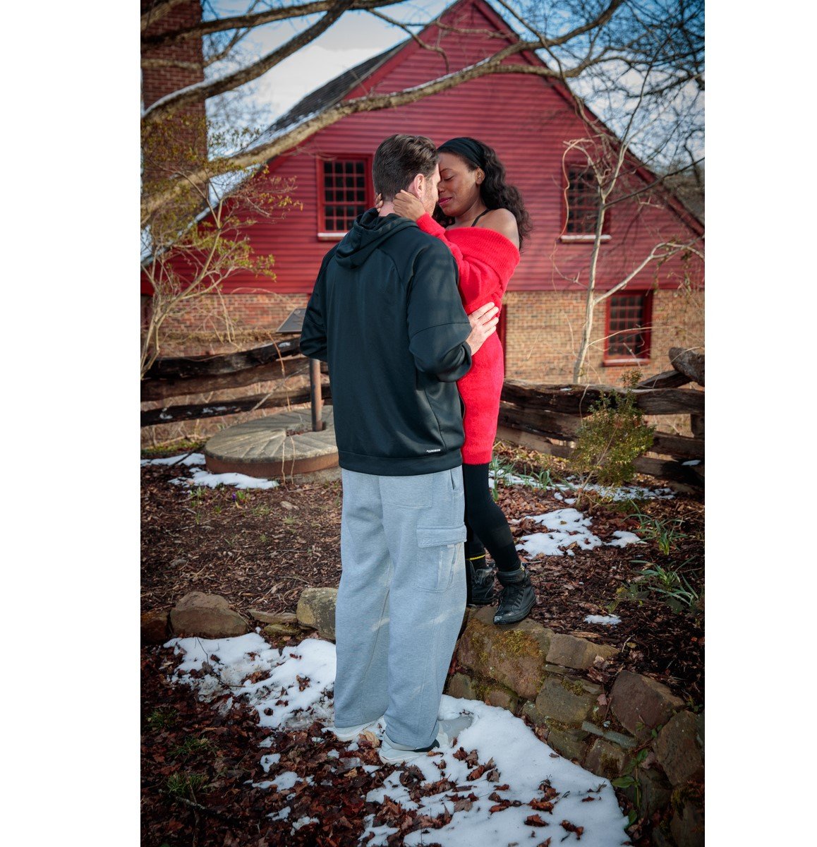 A couple embracing near a red house with snow on the ground, surrounded by a wooden fence and trees.