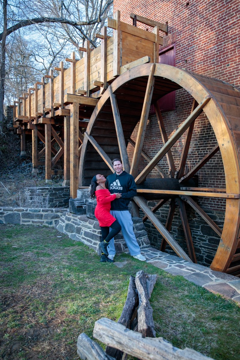 Two people standing by a large wooden water wheel attached to a brick building, with trees and a grassy area around them.