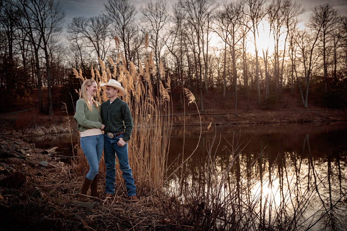 A couple standing by a lake with tall grasses at sunset, wearing casual western-style clothing and cowboy hats, amidst a backdrop of bare trees reflecting in the water.