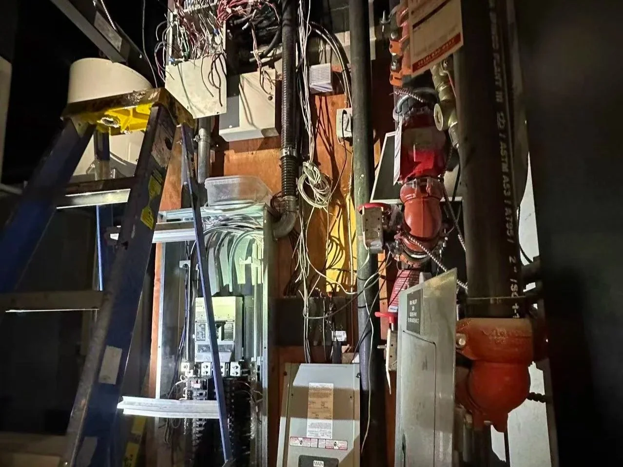 Interior view of an electrical or mechanical room with a ladder, wiring, control panels, pipes, and equipment.