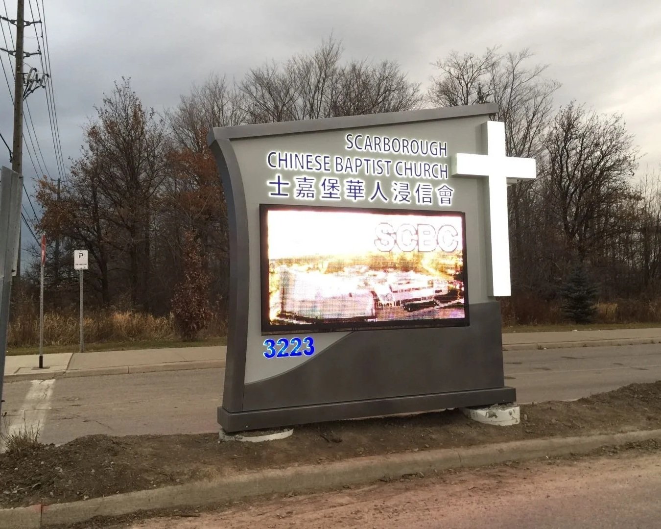 Sign for Scarborough Chinese Baptist Church with a cross, displaying the address 3223 and a digital screen showing an image of a parking lot.