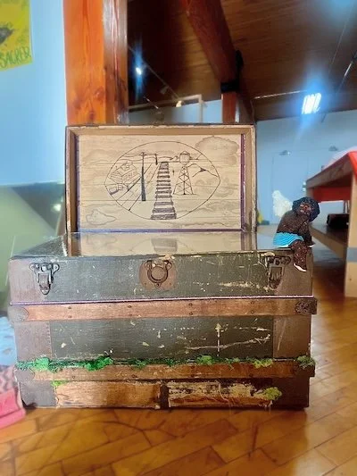 Image of artwork by BakiBakiBaki: "Trunkette." Close-up image of an open, wooden travel trunk made of dark and medium brown wood that is worn and scratched. Seated on the right corner of a trunk is a beaded figure of a cherub with dark skin.