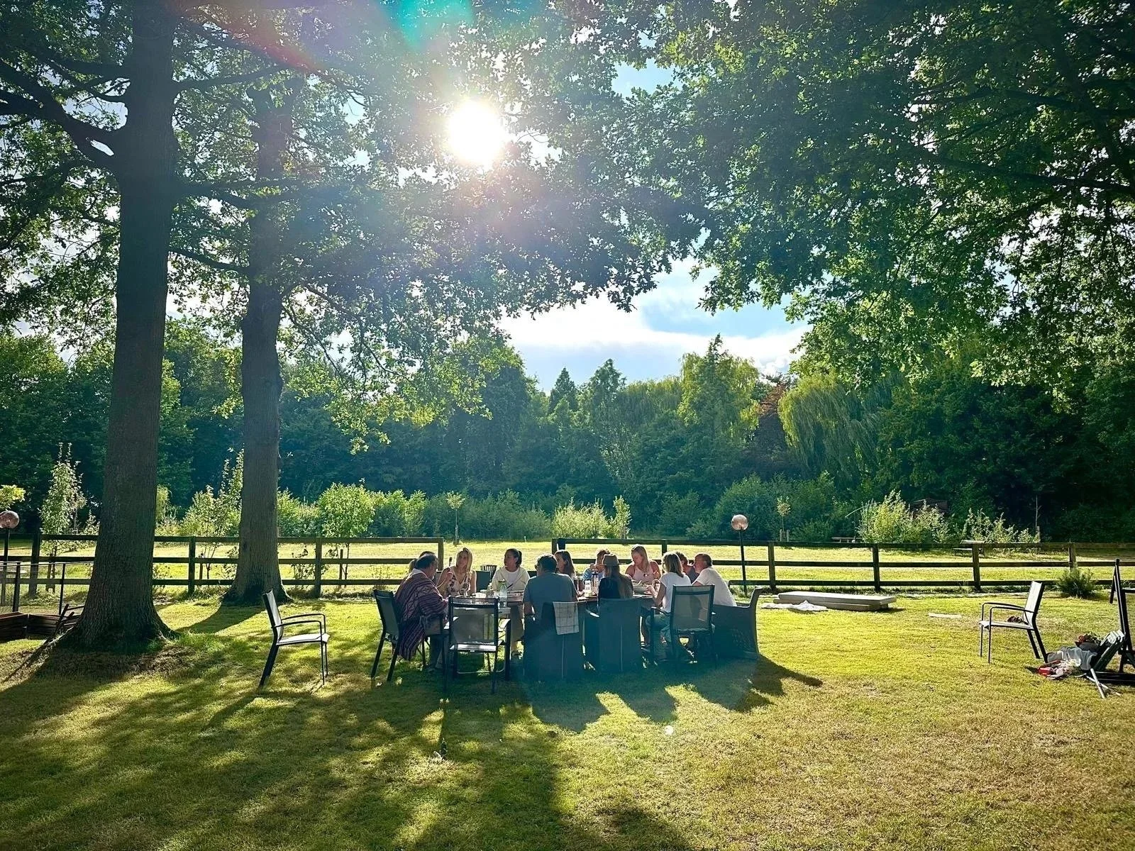 A group of people sitting around a picnic table outdoors on a sunny day, with trees and a wooden fence in the background.