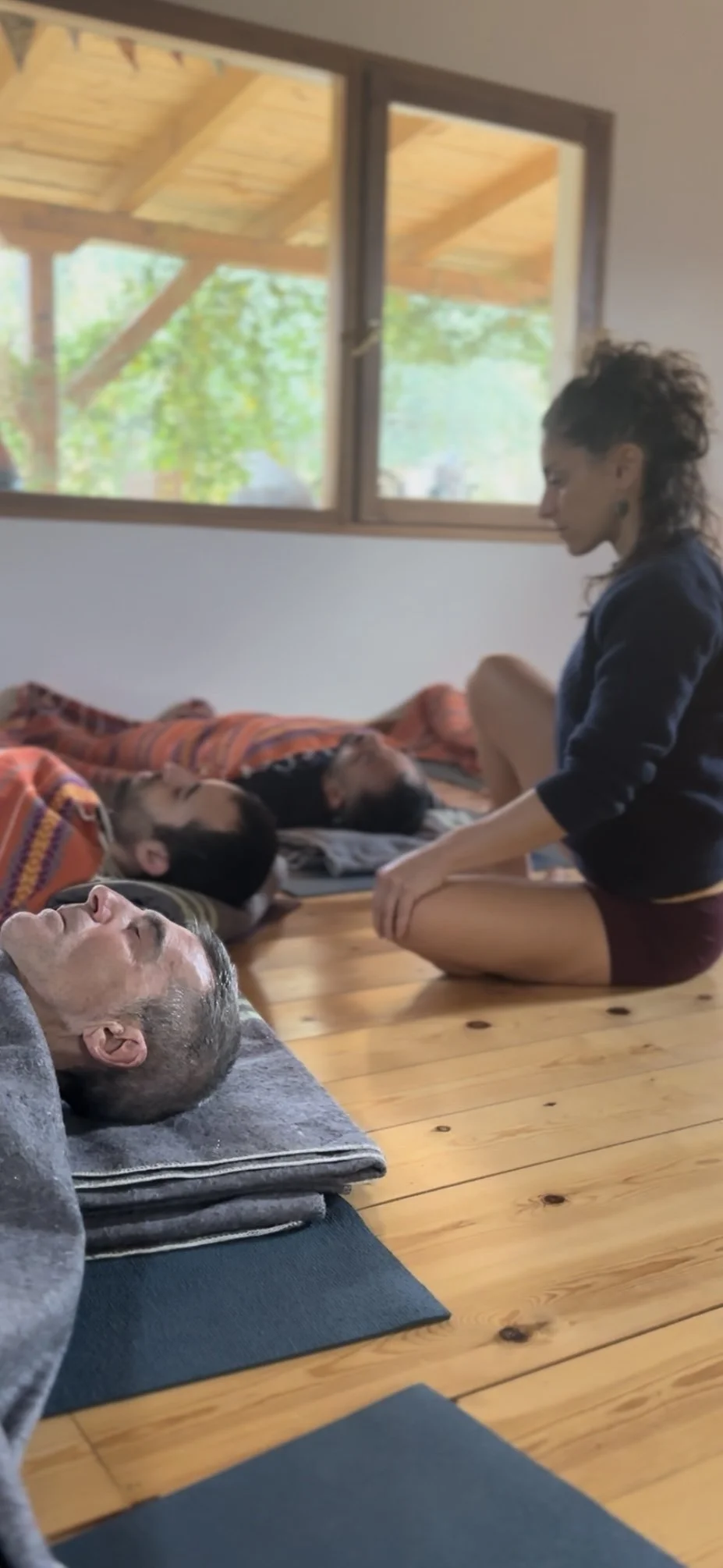 A woman and a man practicing yoga in a room with wooden floors and large windows. Two people are lying on their backs on mats, while the woman sits cross-legged between them, engaging in meditation or a gentle yoga pose.