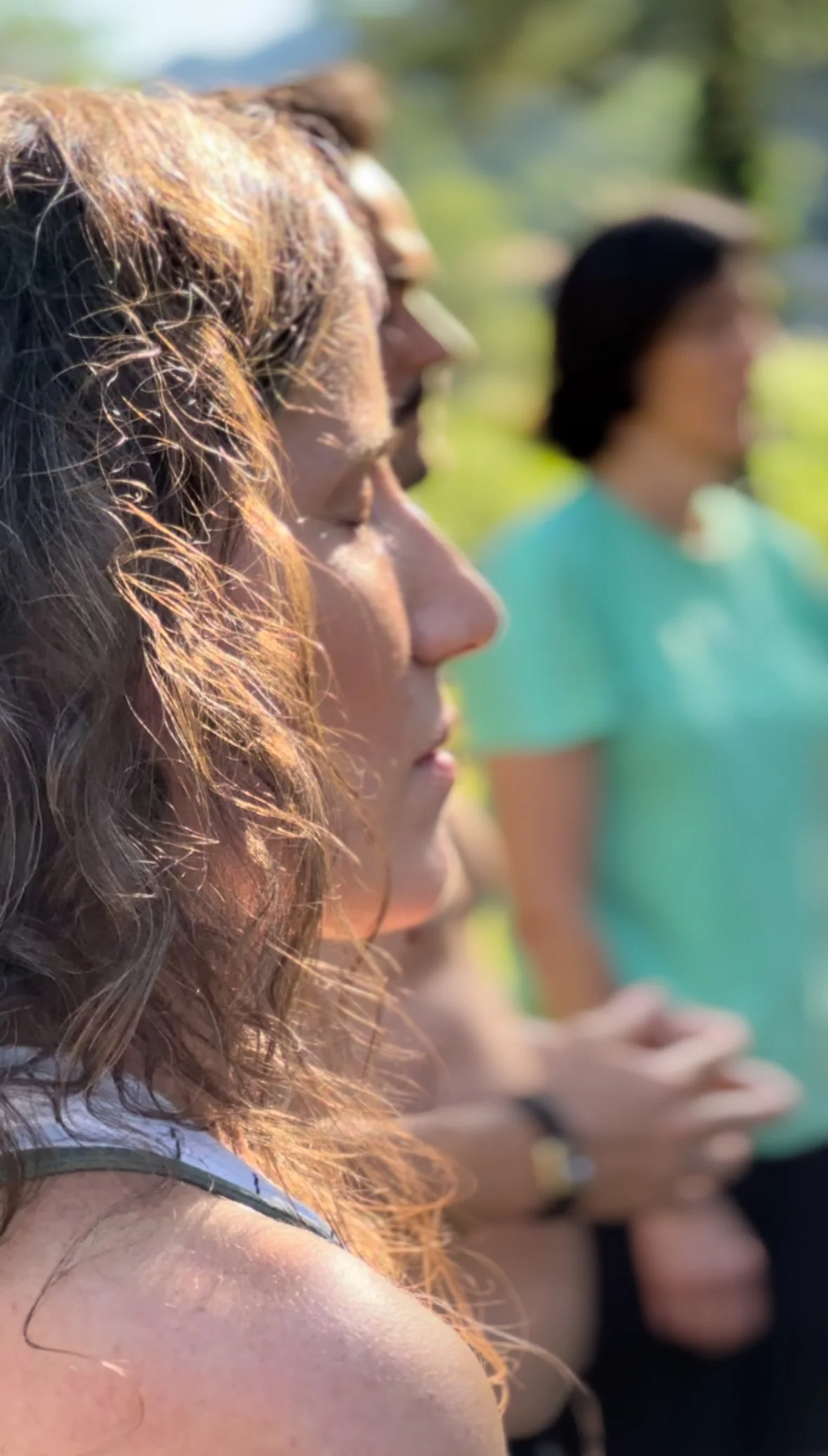 Close-up of a woman with curly brown hair in profile, outdoors on a sunny day, with other people blurred in the background.
