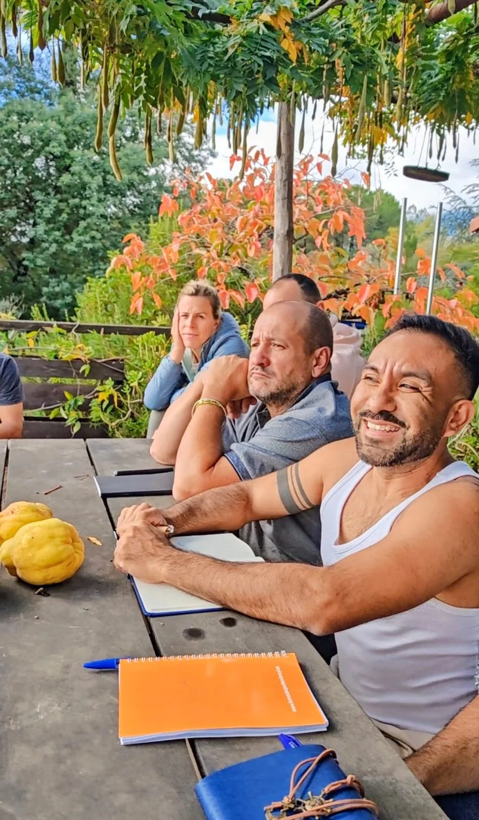 People sitting at a wooden outdoor table with notebooks and pens, surrounded by green and orange foliage. There are pumpkins on the table.