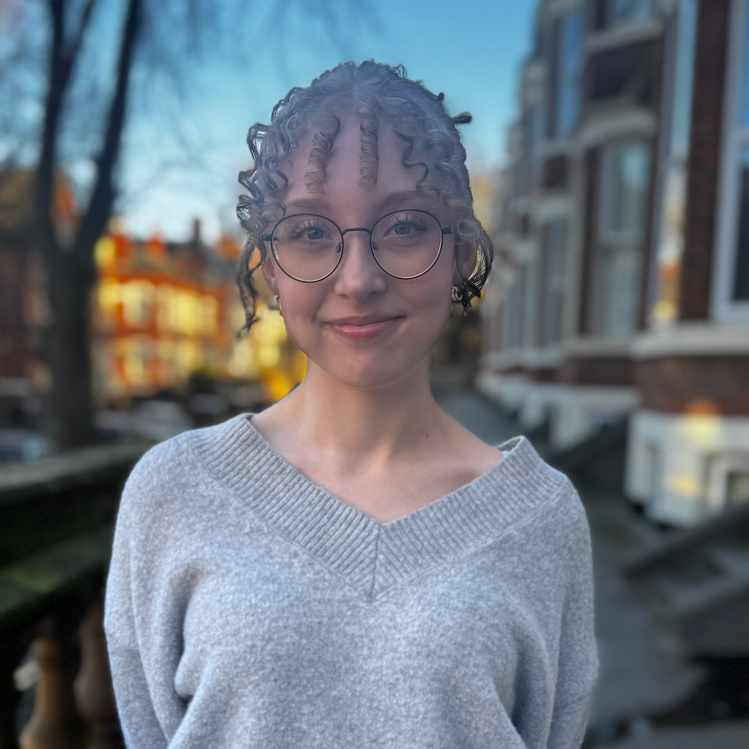 A smiling young woman with curly hair and glasses standing outdoors on a sidewalk in front of residential buildings during sunset.