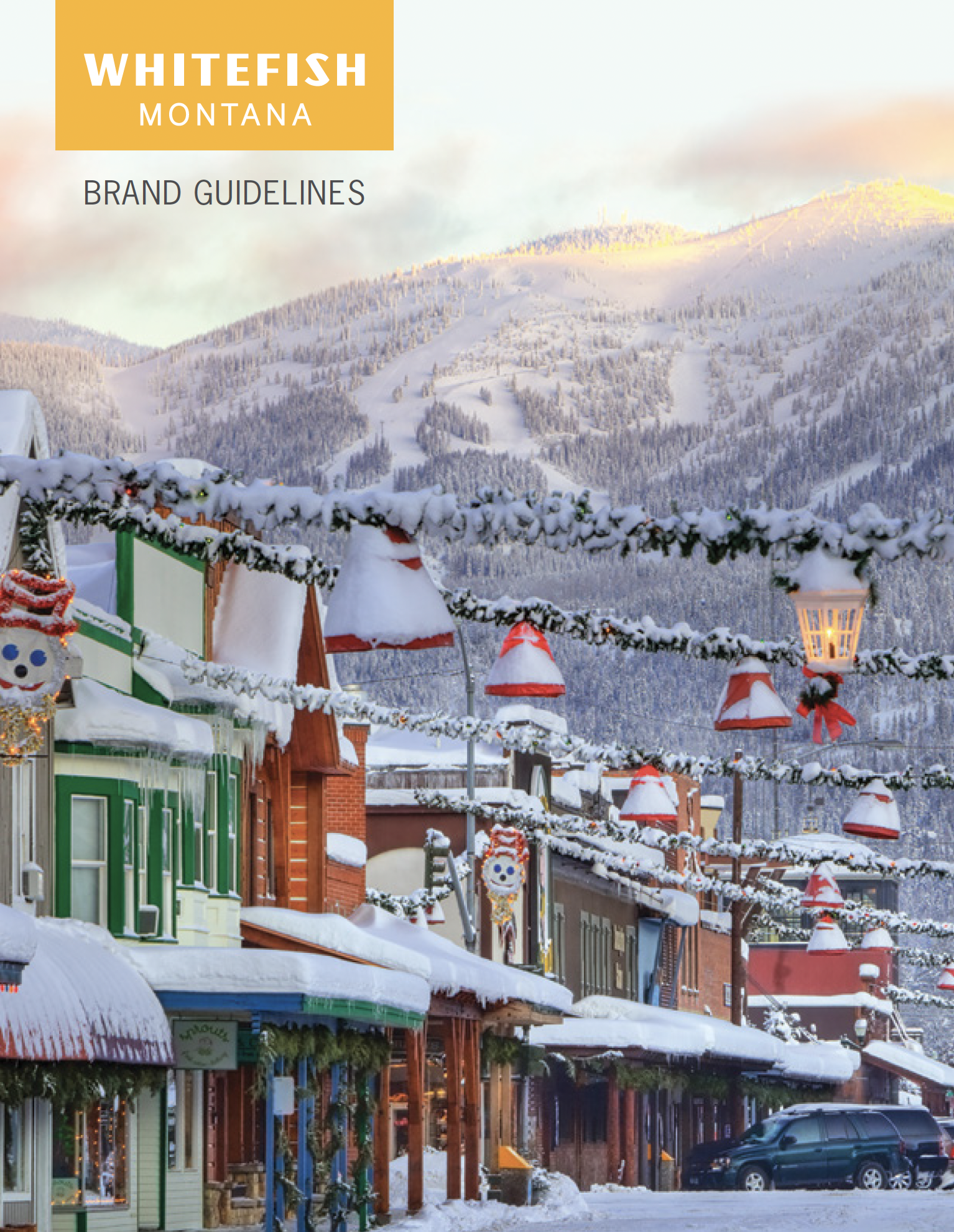 Snow-covered storefronts on a street with decorative holiday lights and ornaments, with a mountain in the background during winter.