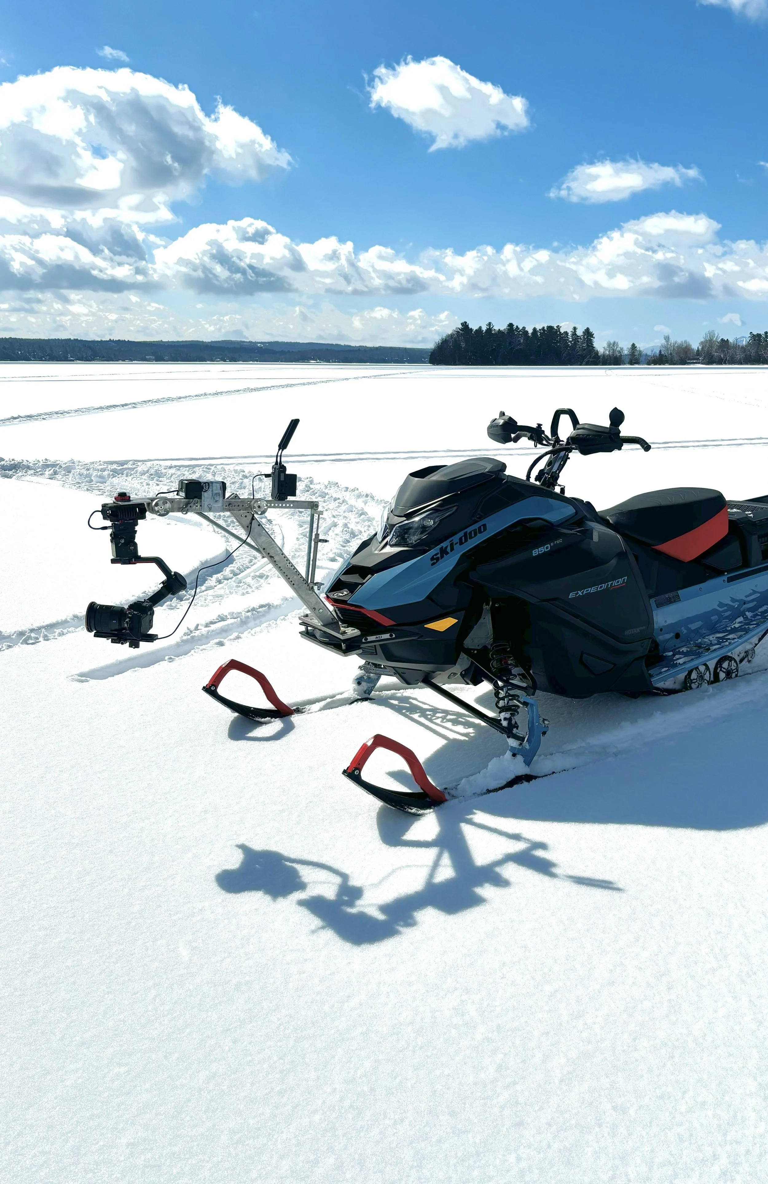 A snowmobile on a snow-covered field with an icy lake and a tree line in the distance, under a bright blue sky with scattered clouds.
