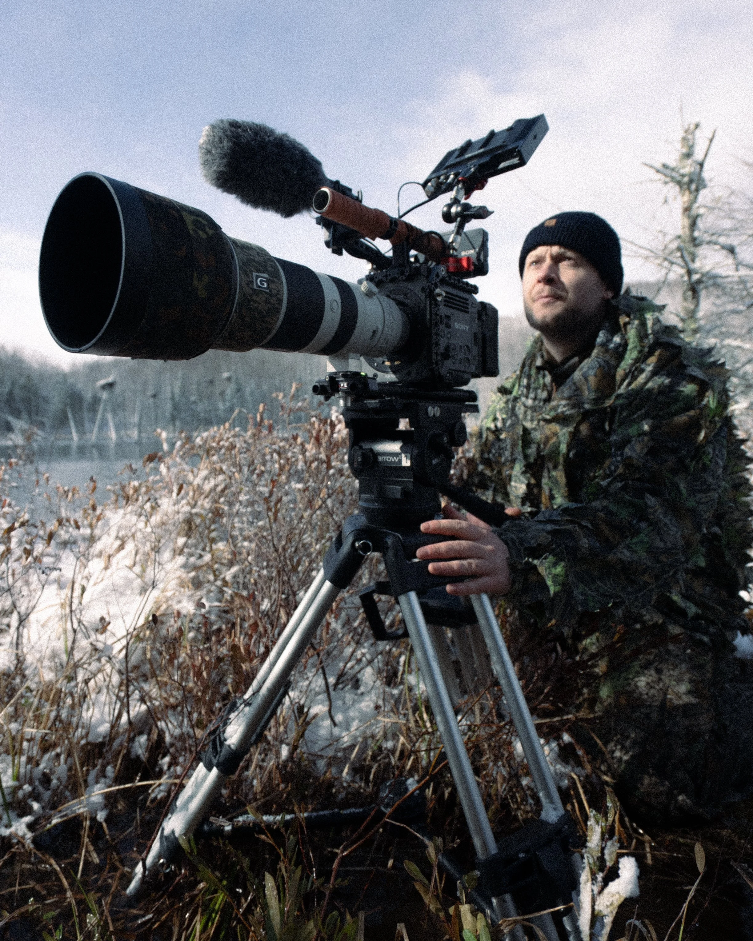 A person in camouflage outdoor clothing and a black beanie looks into the distance while operating a large camera with a yellow and black lens, mounted on a tripod, in a snowy outdoor setting with leafless trees and snow-covered ground.
