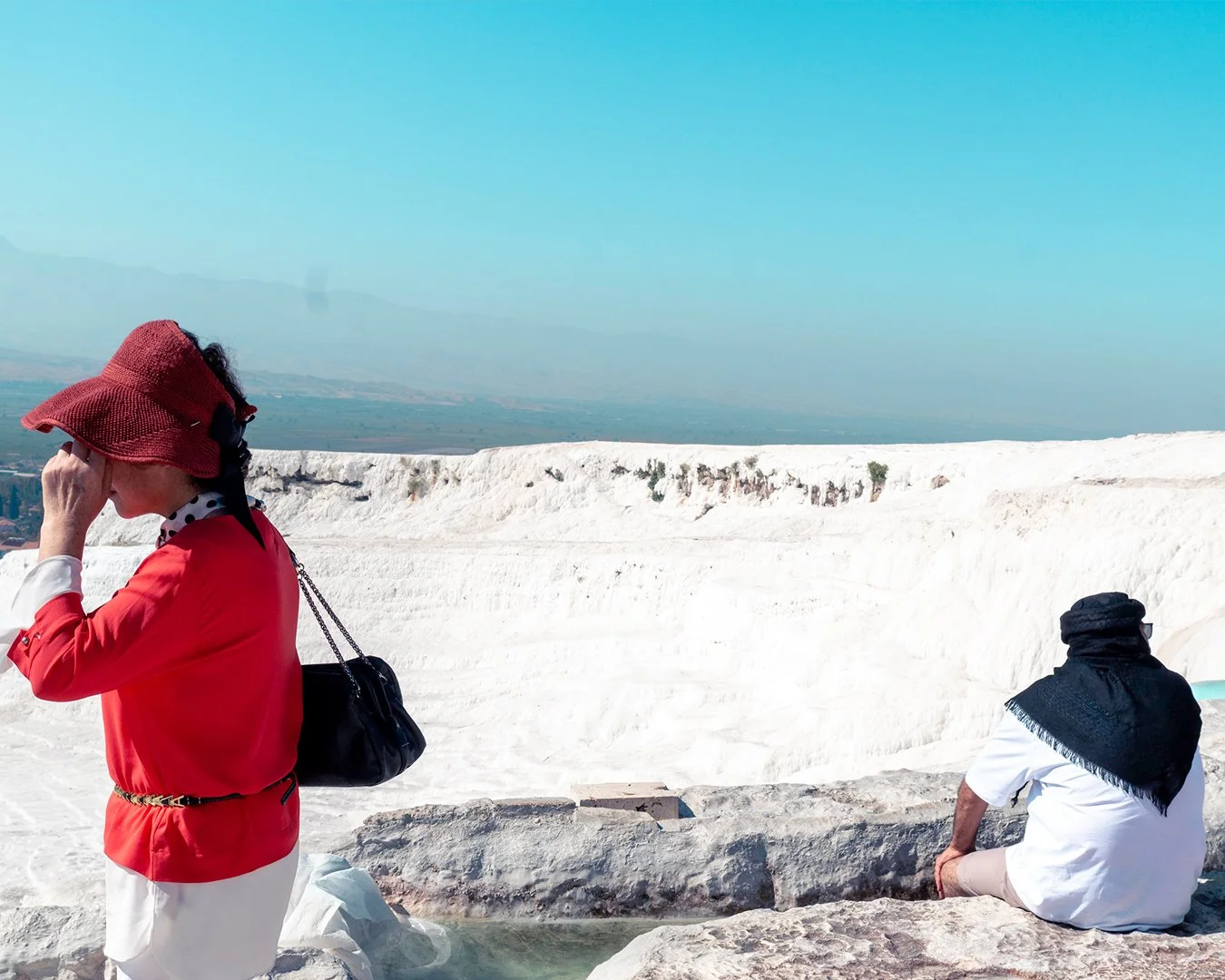 Two tourists, a woman in a red hat and dress, and a man with a black head covering, are sitting on rocks near white travertine terraces at Pamukkale, Turkey, with clear blue sky overhead.