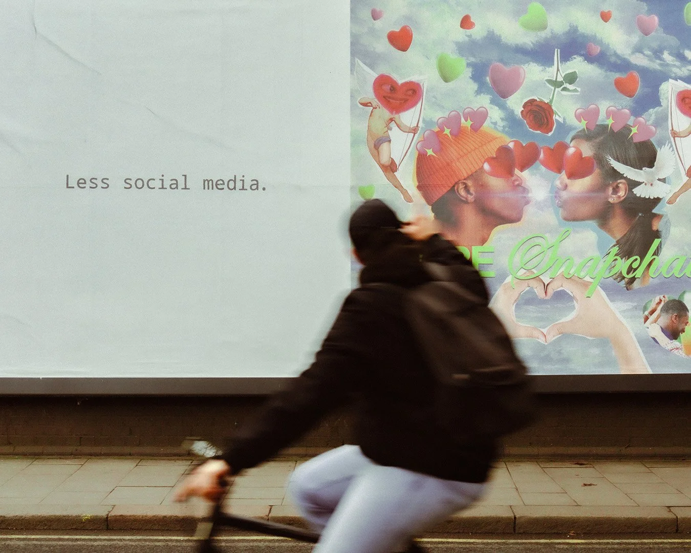 A person riding a bicycle in front of a large poster with the words 'Less social media' on the left side and vibrant, artistic illustrations of two people facing each other, surrounded by hearts, clouds, and other whimsical elements on the right side