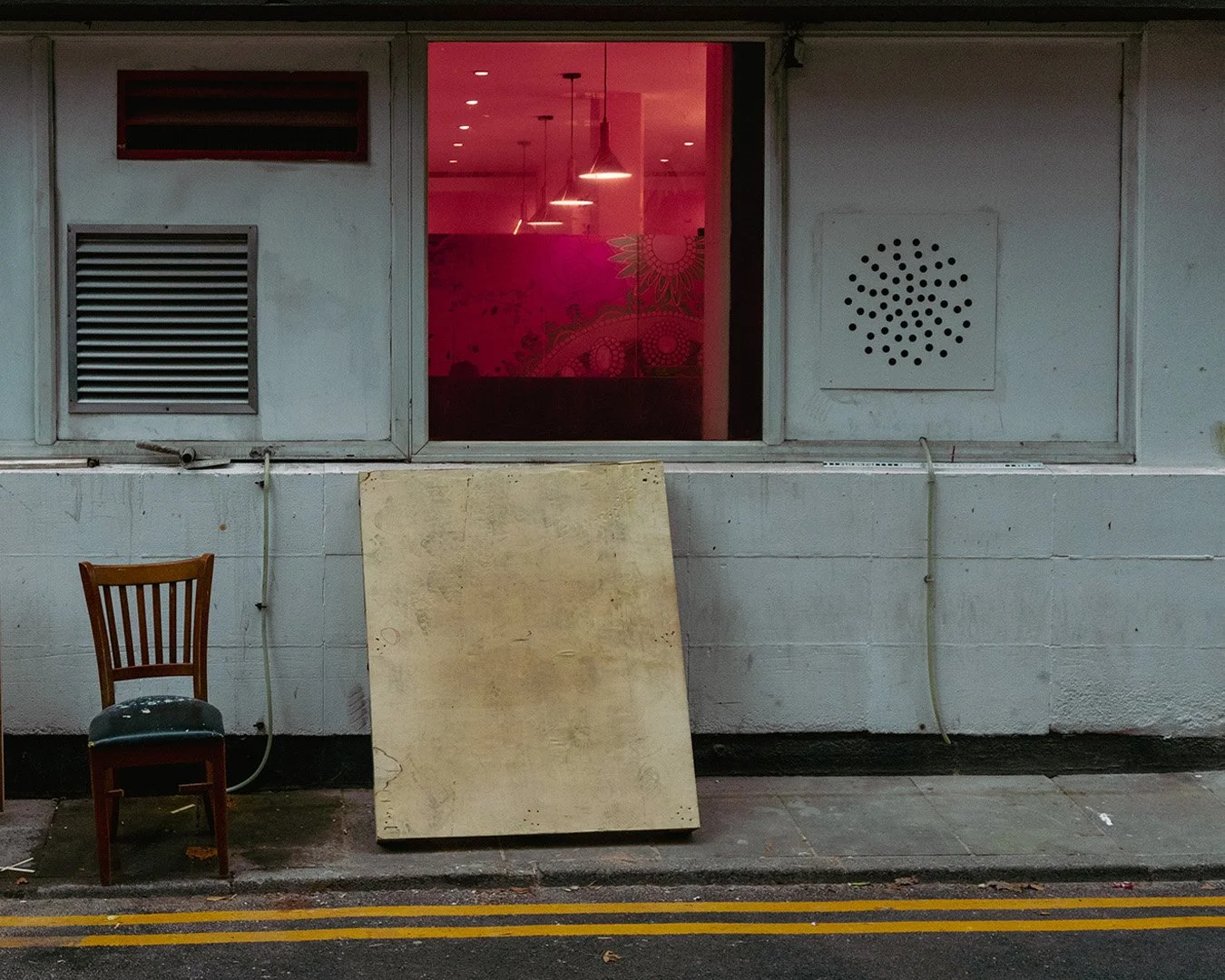 Urban street scene with a worn chair, a large plywood board, and a building with a window showing a pink-lit interior.