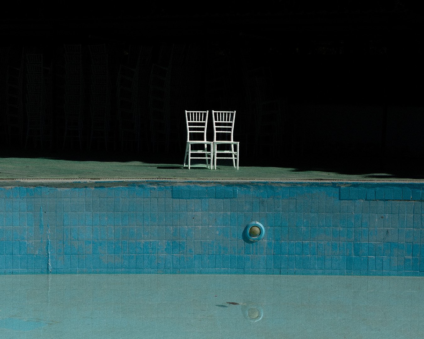 Two white chairs placed side by side on green ground behind a blue swimming pool, with a black background and faint stacked chairs in the distance.