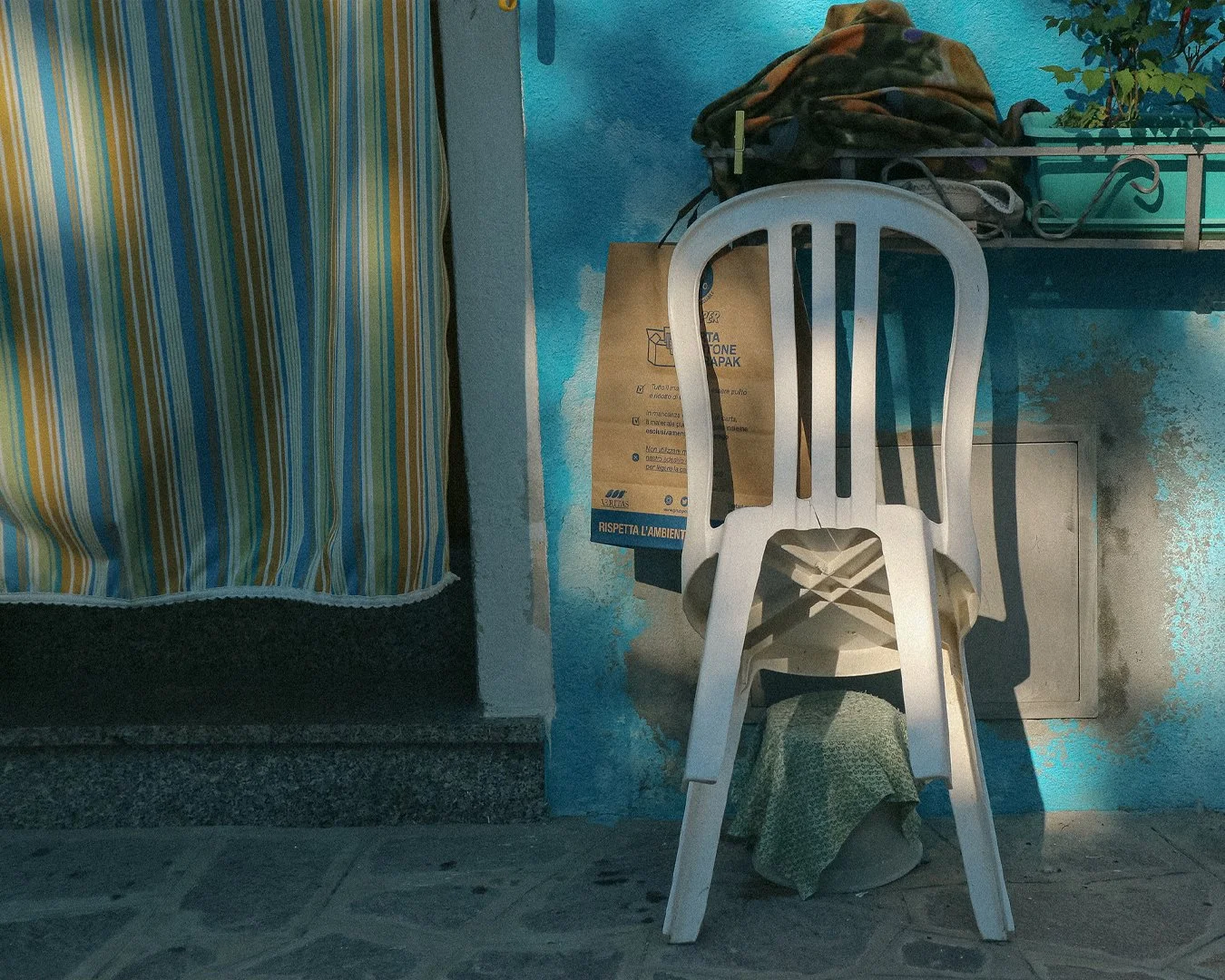 A white plastic chair stacked upside down on a brown bag, with a cloth underneath, against a blue wall with signs and a shelf with a pile of clothes or blankets, next to a window with yellow and green striped curtain.