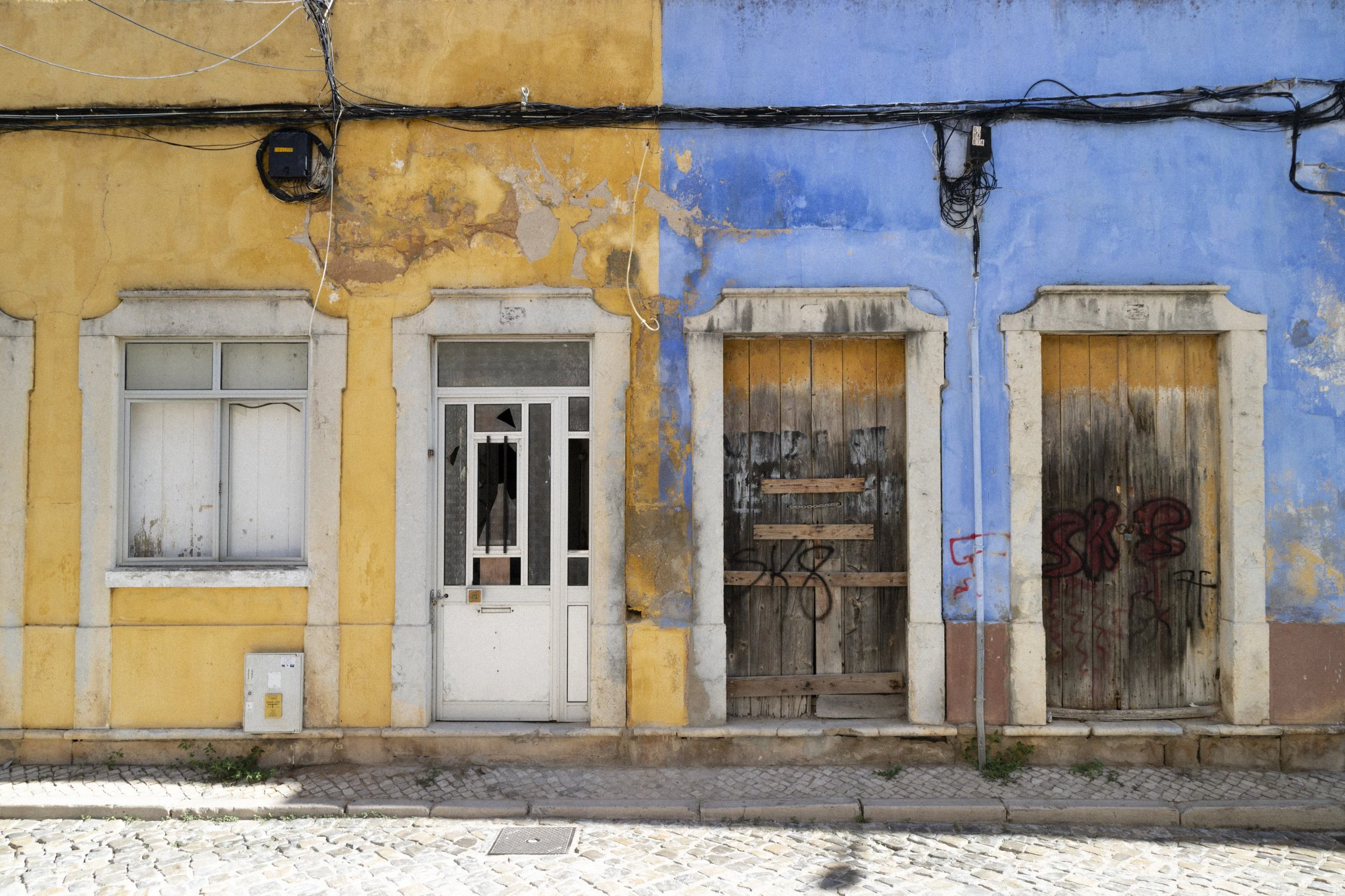 An old building with a yellow and blue painted facade, showing peeling paint, two closed boarded-up doors, a window with barred glass, exposed electrical wires, and graffiti.