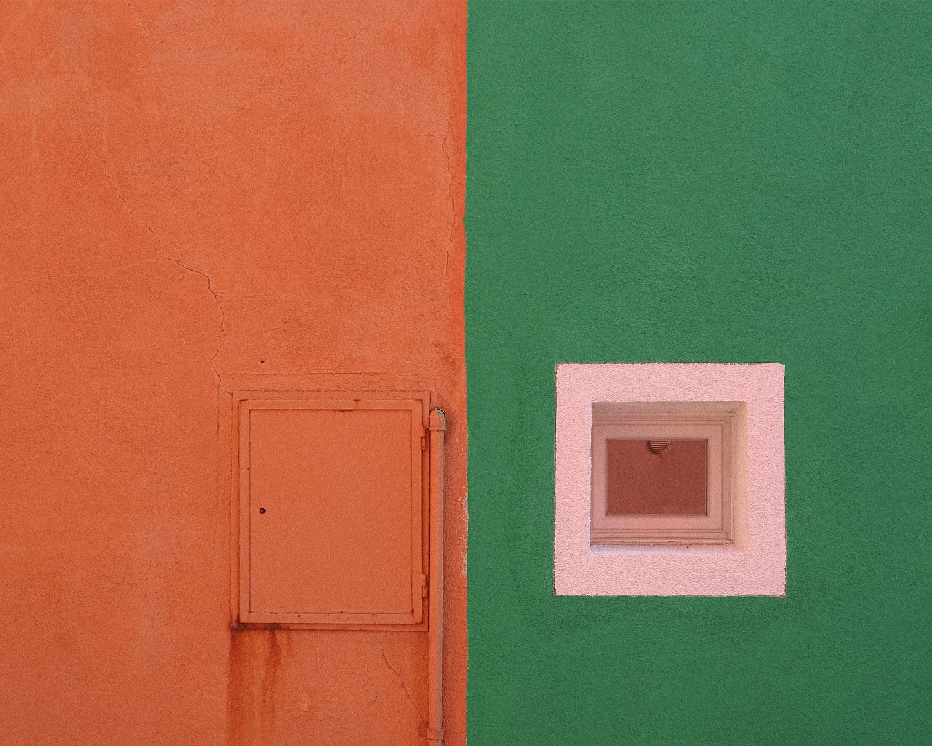A wall split into two colors, orange on the left with a cracked texture and a small closed door, and green on the right with a square window framed in white.