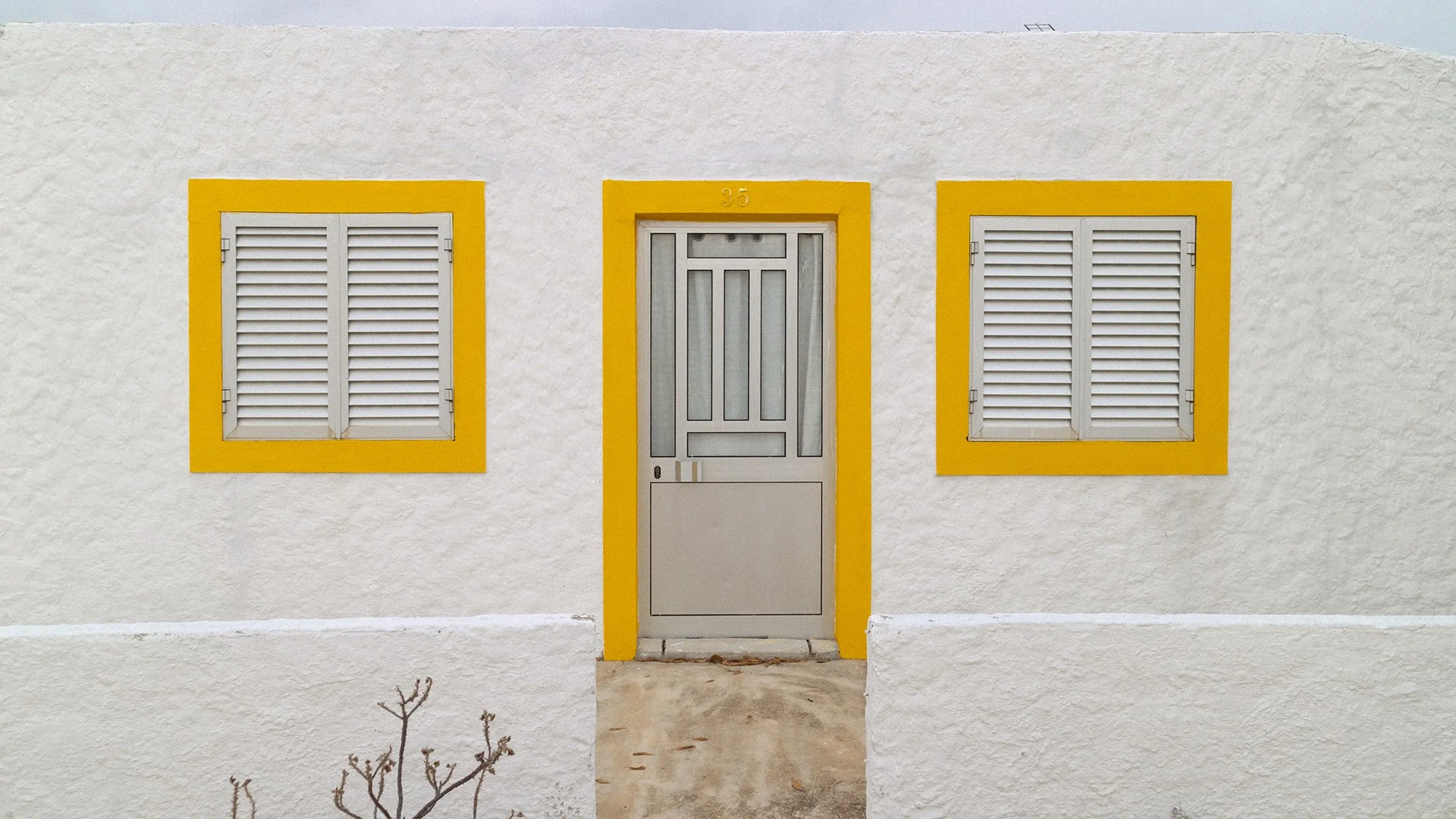 A white stucco house with yellow trim around a door and two windows. The door has a window and a lock, and the house number 35 is above the door. The two windows have white shutters. There is a concrete walkway in front of the house.