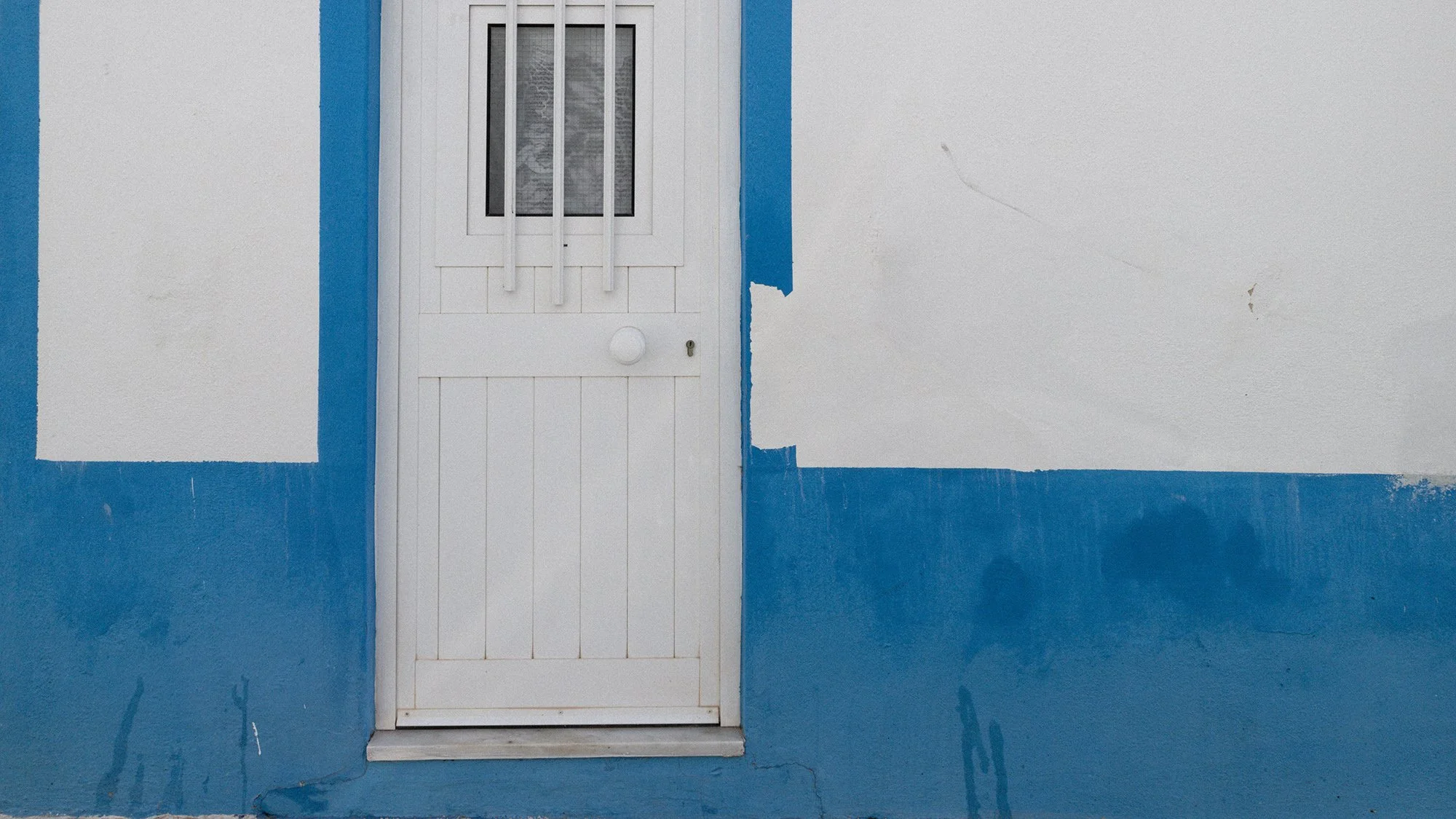 White door with a small window and security bars, set in a wall painted white and blue, with the blue on the lower portion and around the edges of the door.