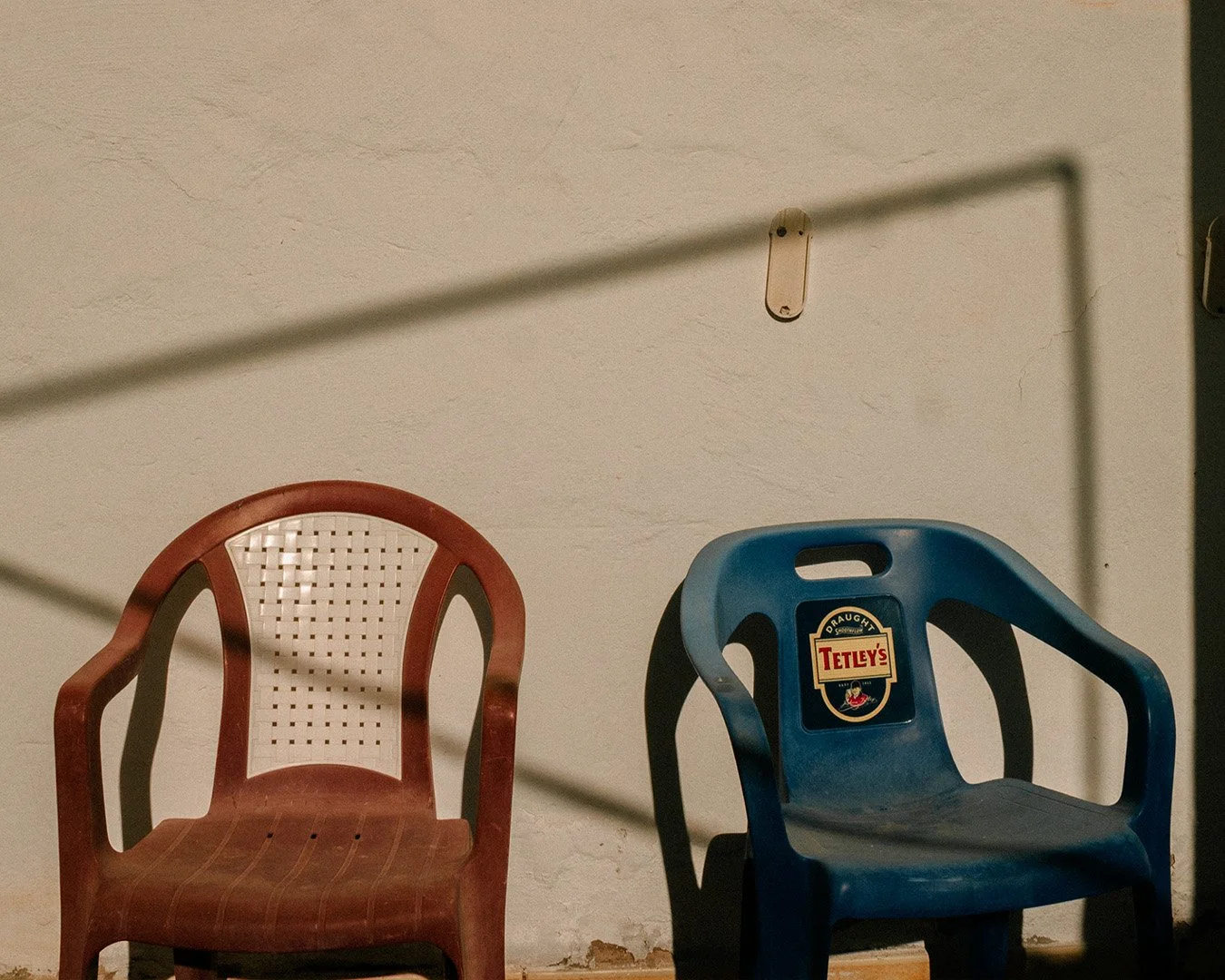 Two plastic chairs, one red with beige backrest and one blue with a Tetley's beer logo, in front of a plain white wall with a floating shelf.