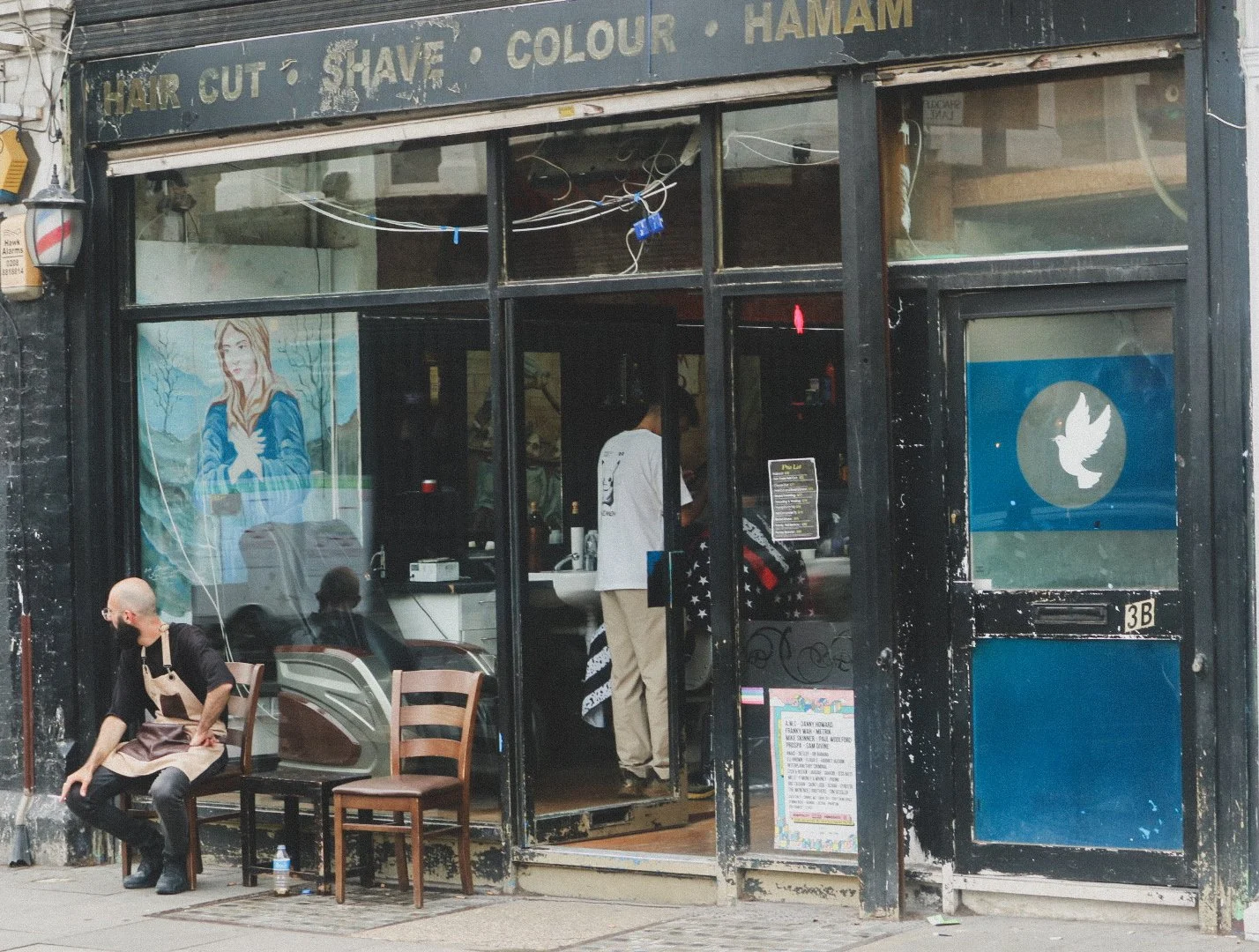 A barbershop exterior with a faded sign listing services like haircuts, shaves, coloring, and hamam. Two chairs outside with a bald man sitting on one, wearing an apron. Through the large front window, a person inside with a white shirt is seen from 