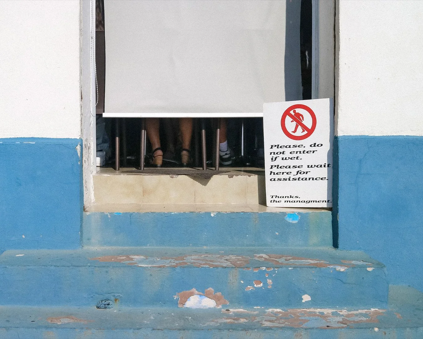 People sitting behind a desk or counter with only their legs visible, seen through a small opening in a wall. A sign with a no-entry symbol and text, asking visitors not to enter if wet and to wait for assistance.