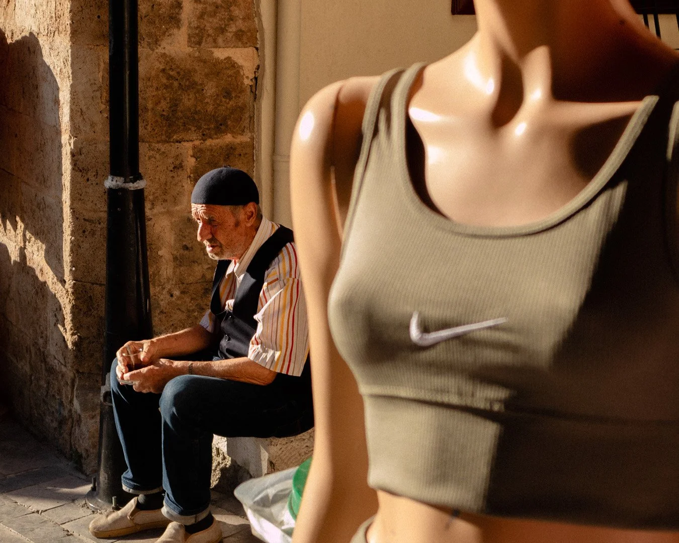 An older man with a beard and hat sitting on a bench against a stone wall, looking at his phone, with a mannequin wearing a green sports bra in the foreground.