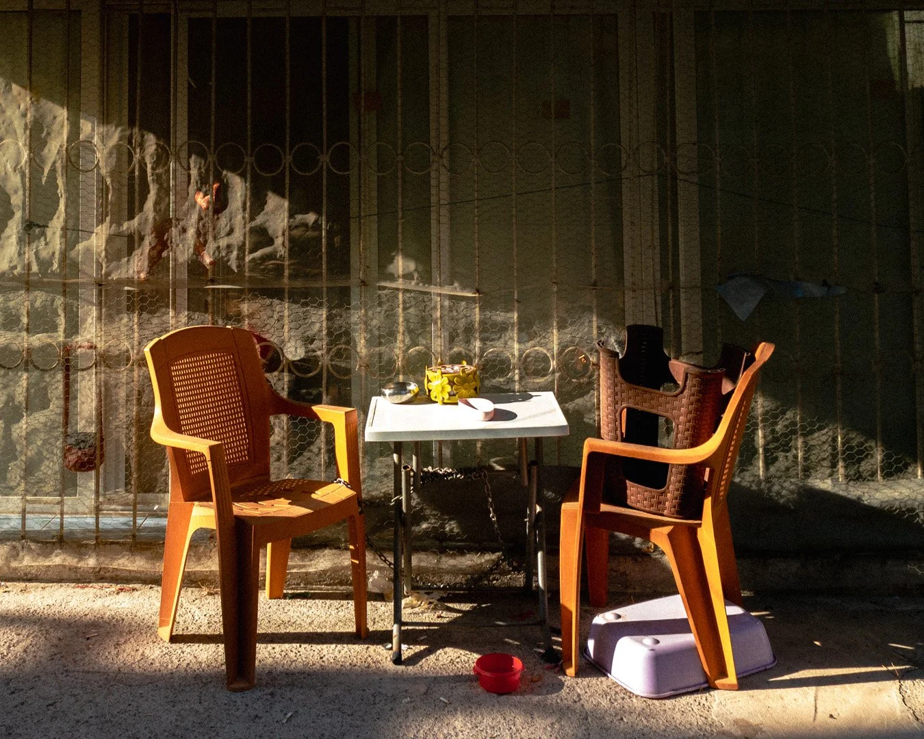 Two plastic chairs, one yellow and one brown, are positioned around a small table outdoors. On the table, there are a yellow flower-shaped pot, a bowl, and a pink bowl. A purple cat litter box is beside the yellow chair, and a red bowl is on the grou